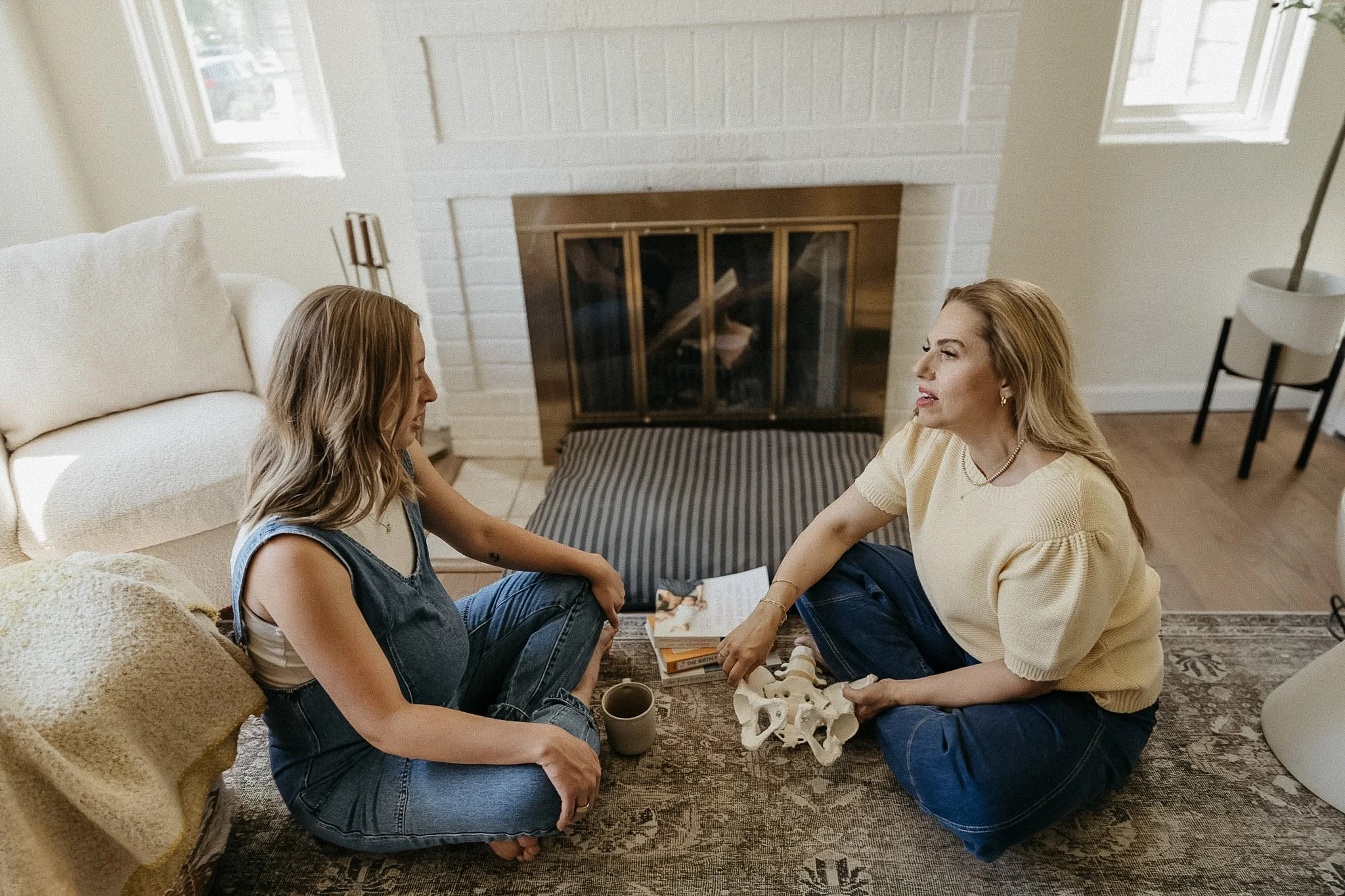 Two women sitting on the floor in a living room, facing each other, engaging in conversation. They are surrounded by books, a mug, and a skeleton model. The room has a fireplace, two windows, and a beige sofa.