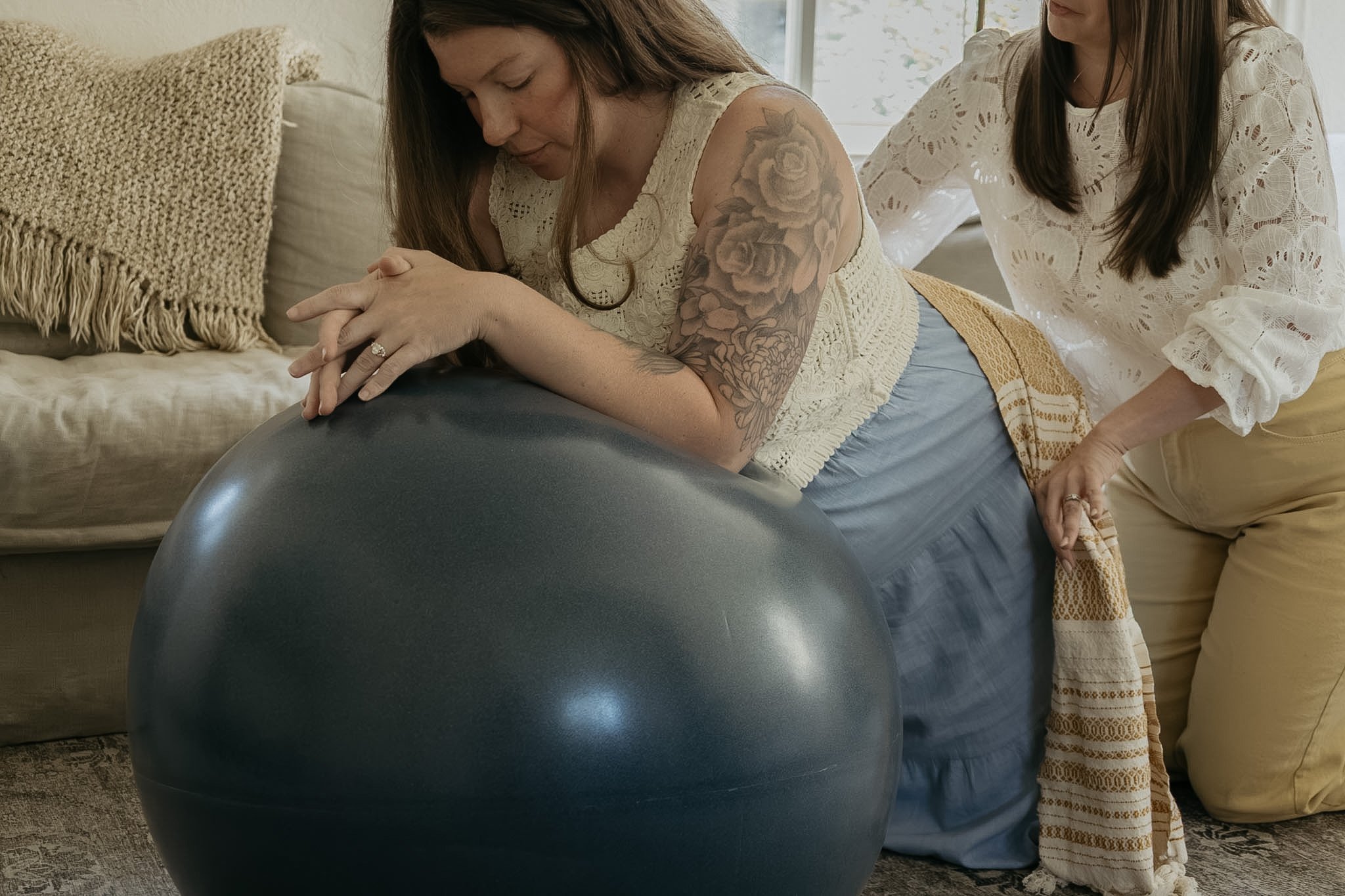 A woman with long brown hair and floral tattoo on her arm doing yoga on a black exercise ball while another woman with brown hair in a white lace shirt assists her in a well-lit room with beige and cream-colored furniture.