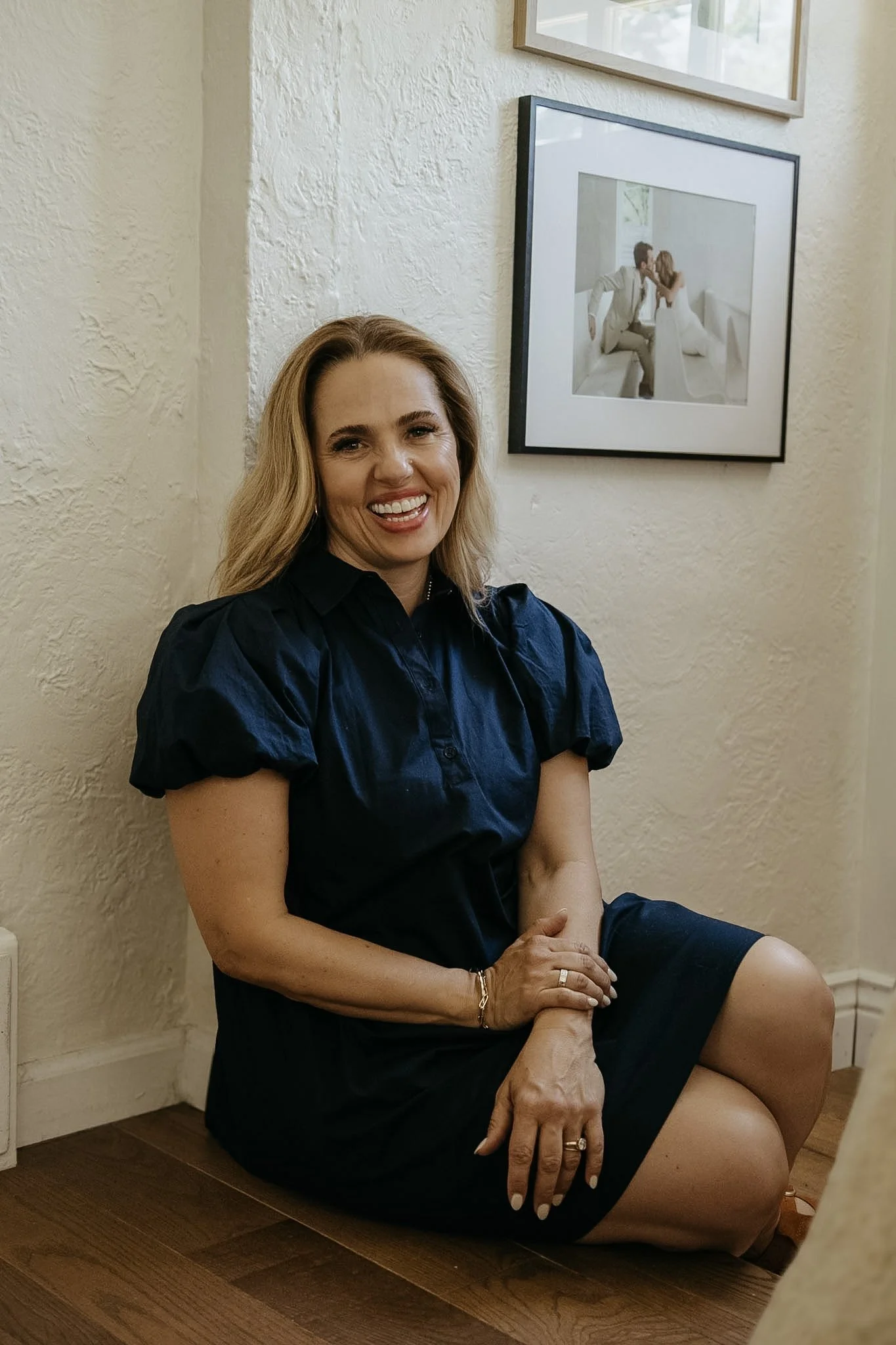 A woman with long blonde hair, wearing a navy blue dress, sitting on the floor beside a wall with framed photos, smiling at the camera.