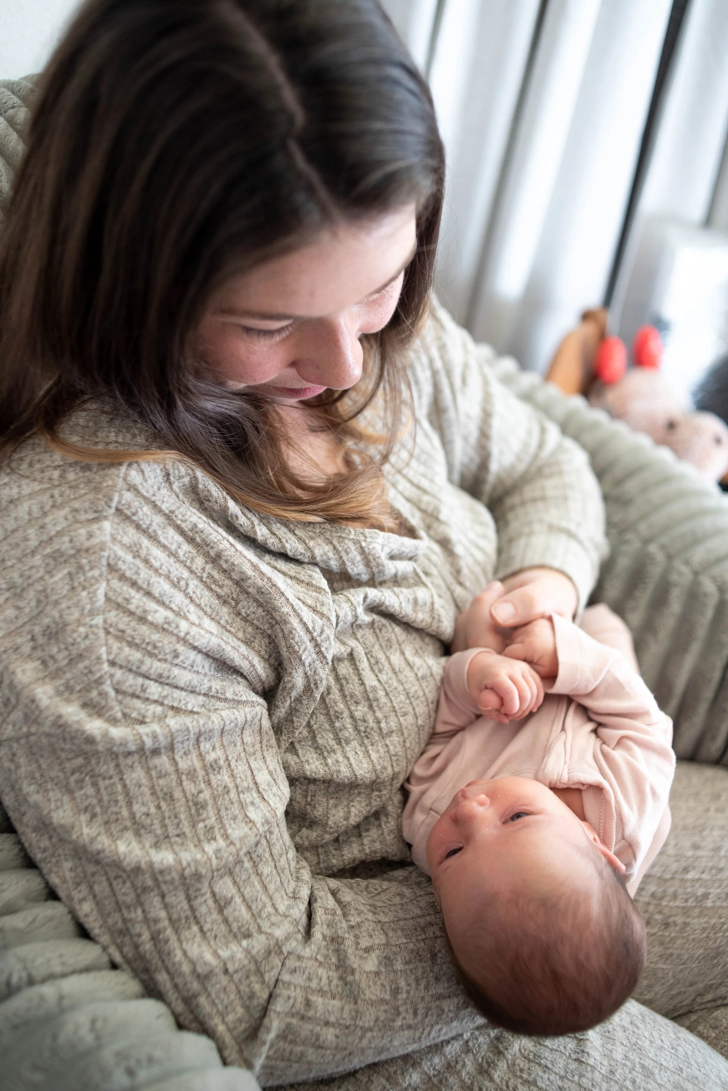 A woman with brown hair looking down lovingly at a baby lying on her lap, inside a room with natural light and a window with curtains in the background.