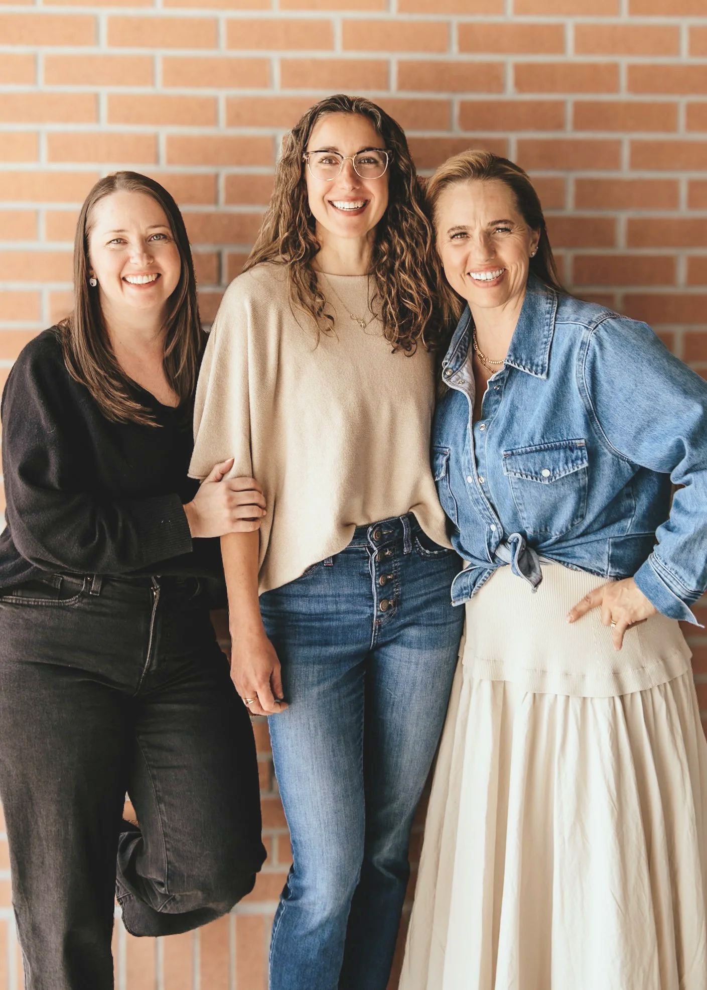 Three women standing close together against a brick wall, smiling at the camera.