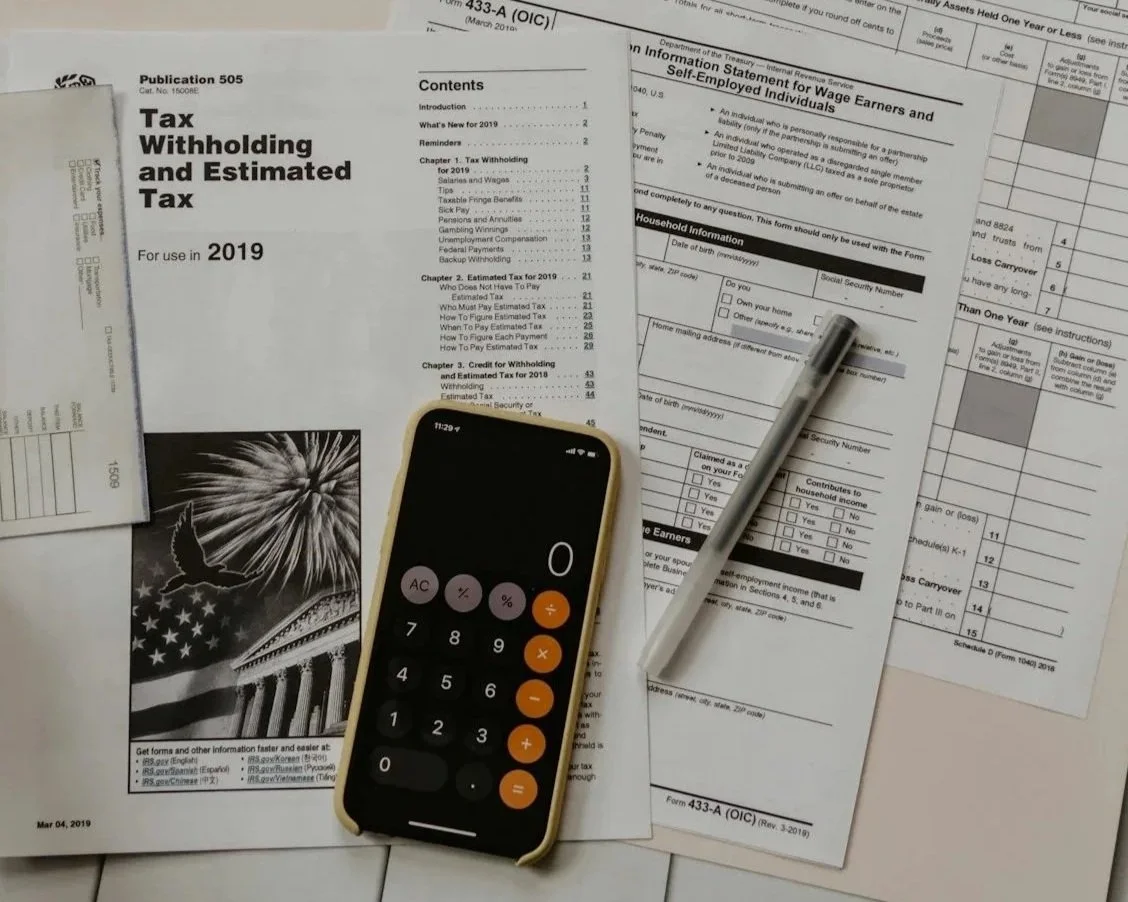 Documentos fiscais e formulários de imposto, um telefone com calculadora, uma caneta e uma folha de papel sobre uma superfície.