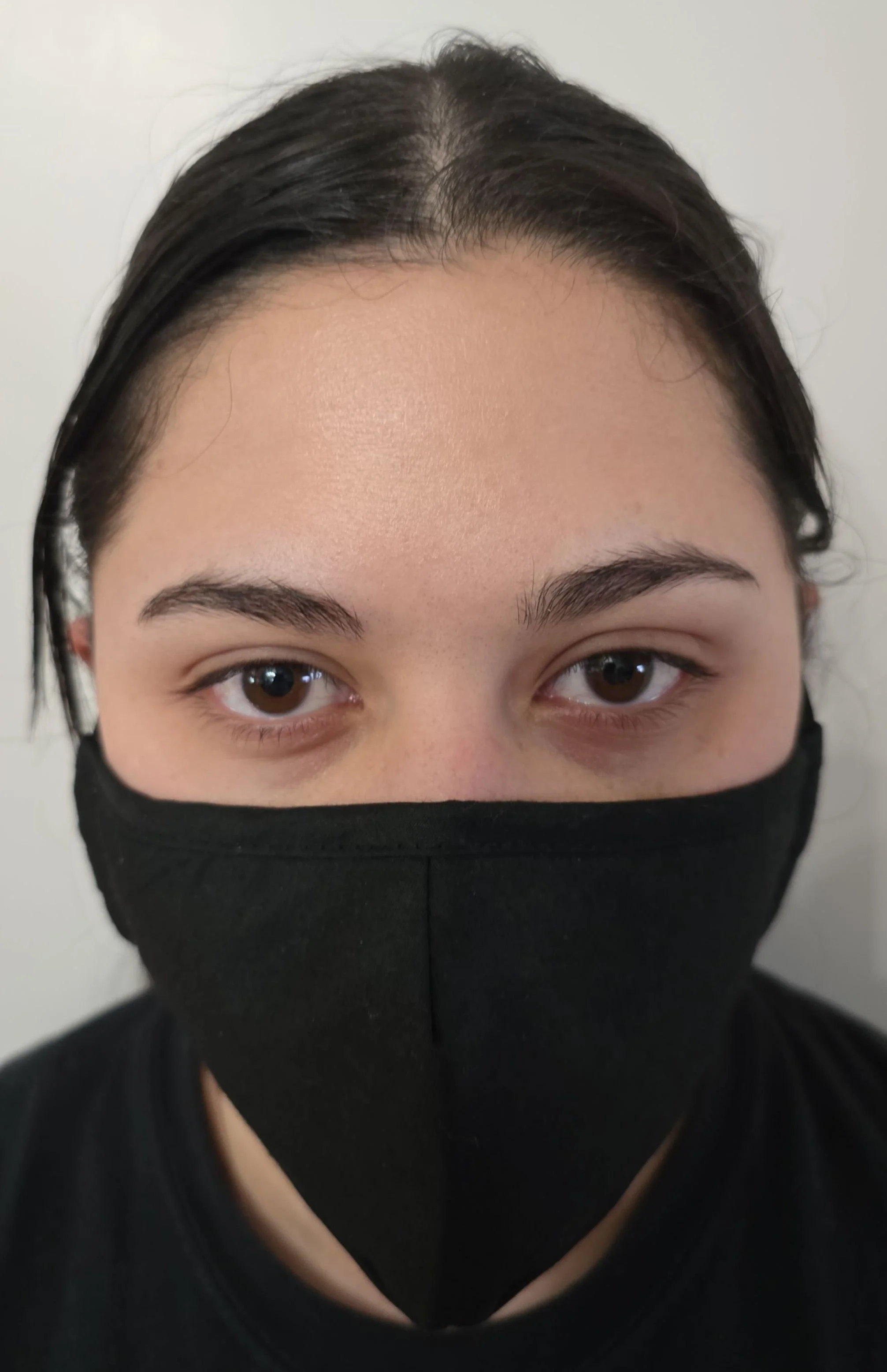 Close-up of a person's face wearing a black face mask, revealing their brown eyes, eyebrows, and dark hair parted in the middle, against a white background.
