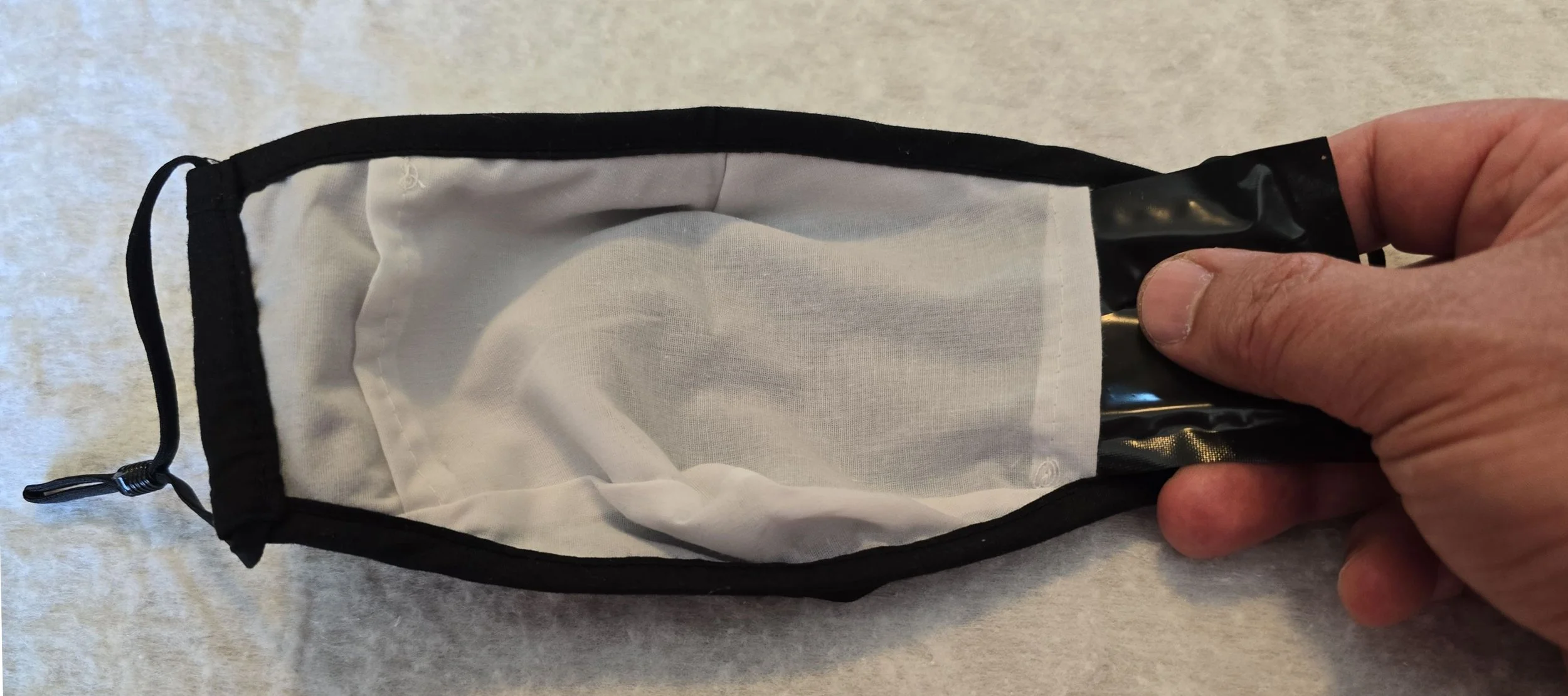 A person holding a black and clear plastic face mask with a white cloth interior, lying on a light-colored surface.