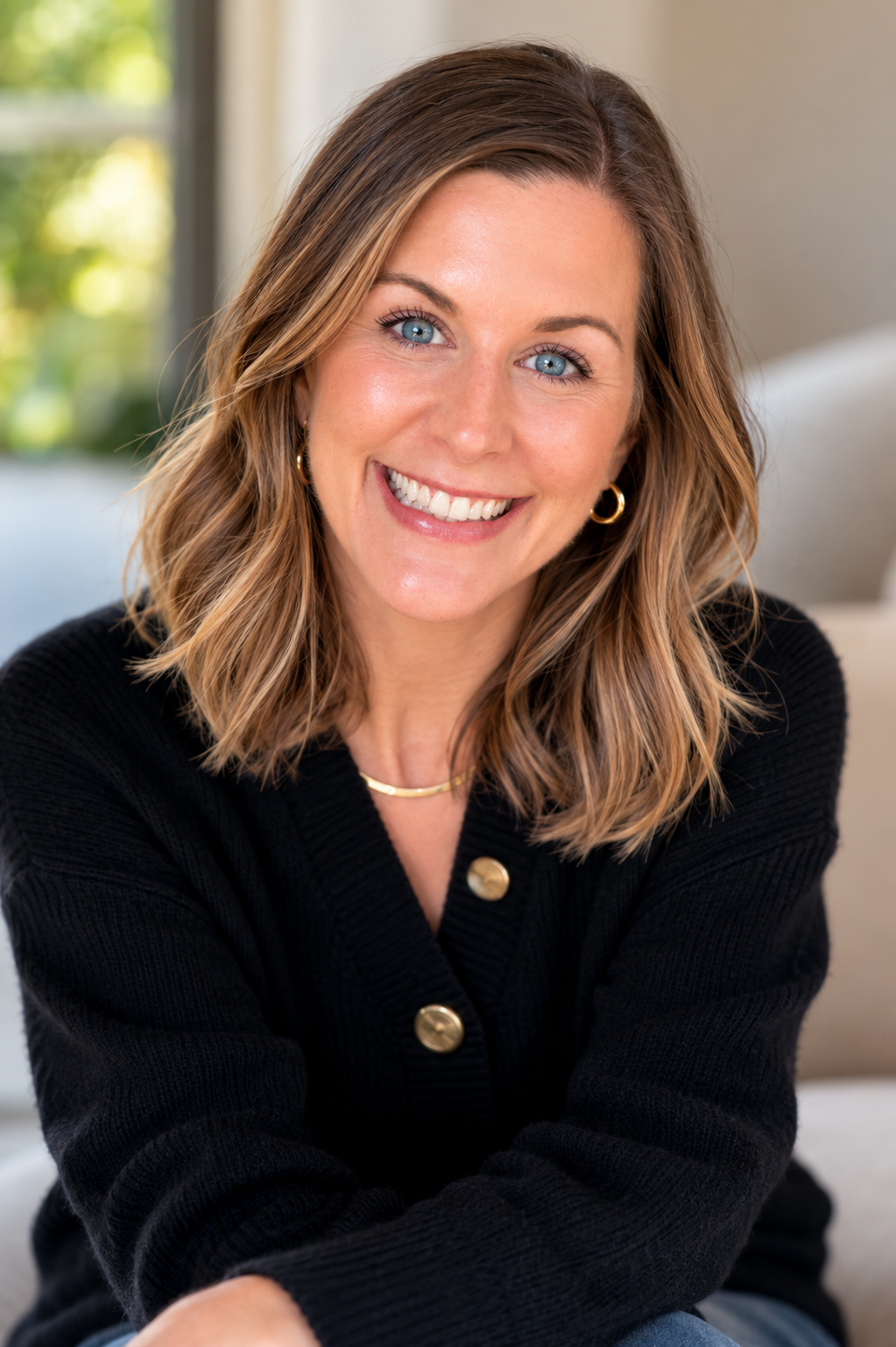A woman with shoulder-length wavy light brown hair and blue eyes, smiling, wearing a black cardigan with gold buttons, gold hoop earrings, and a gold necklace, sitting indoors with a blurred background.