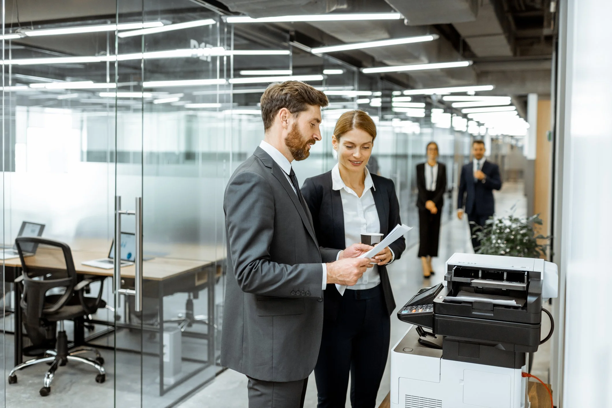 A man and a woman in business attire standing next to a copier in a modern office, looking at documents.