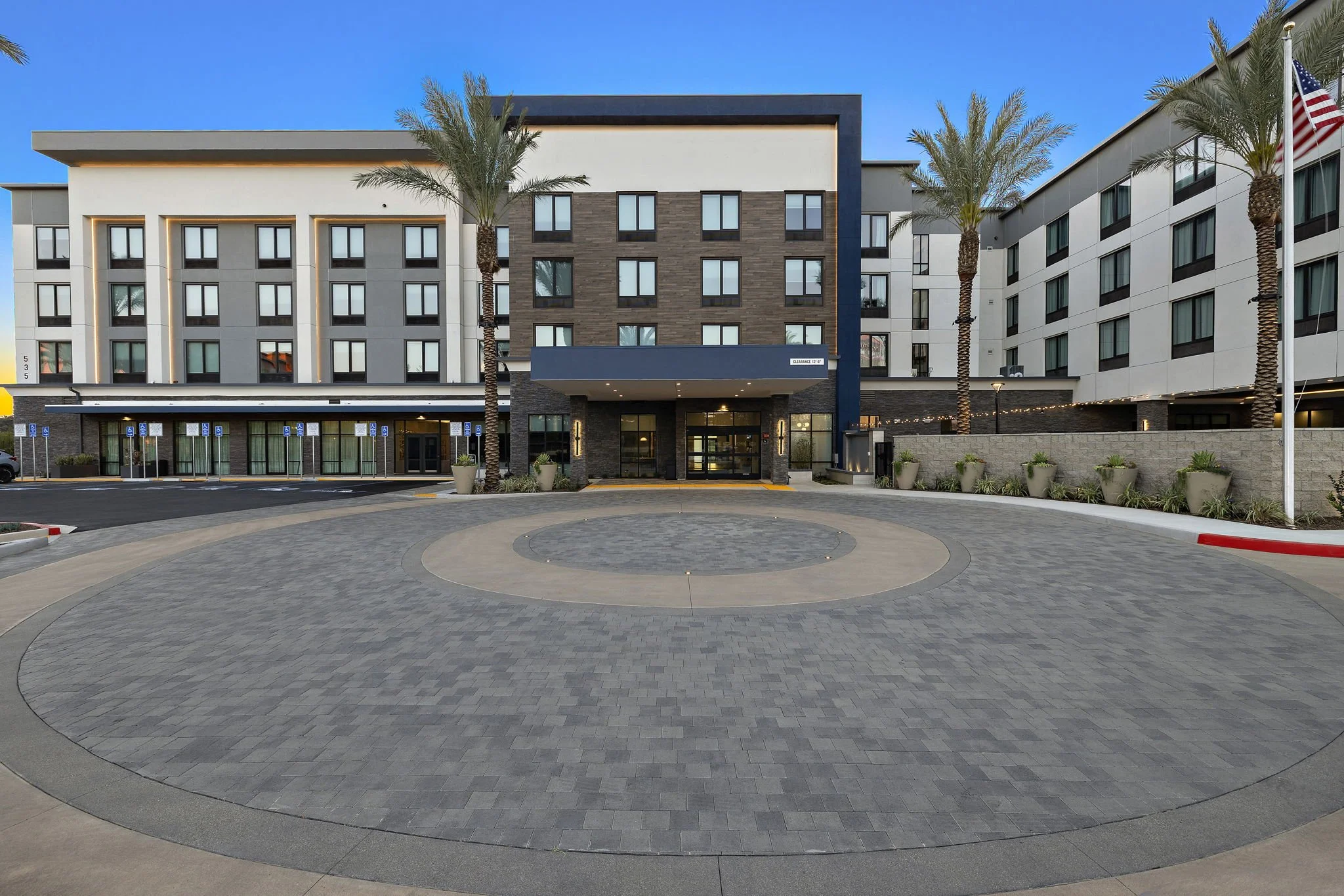 Modern hotel building with palm trees in front, parking spaces, and a clear blue sky.