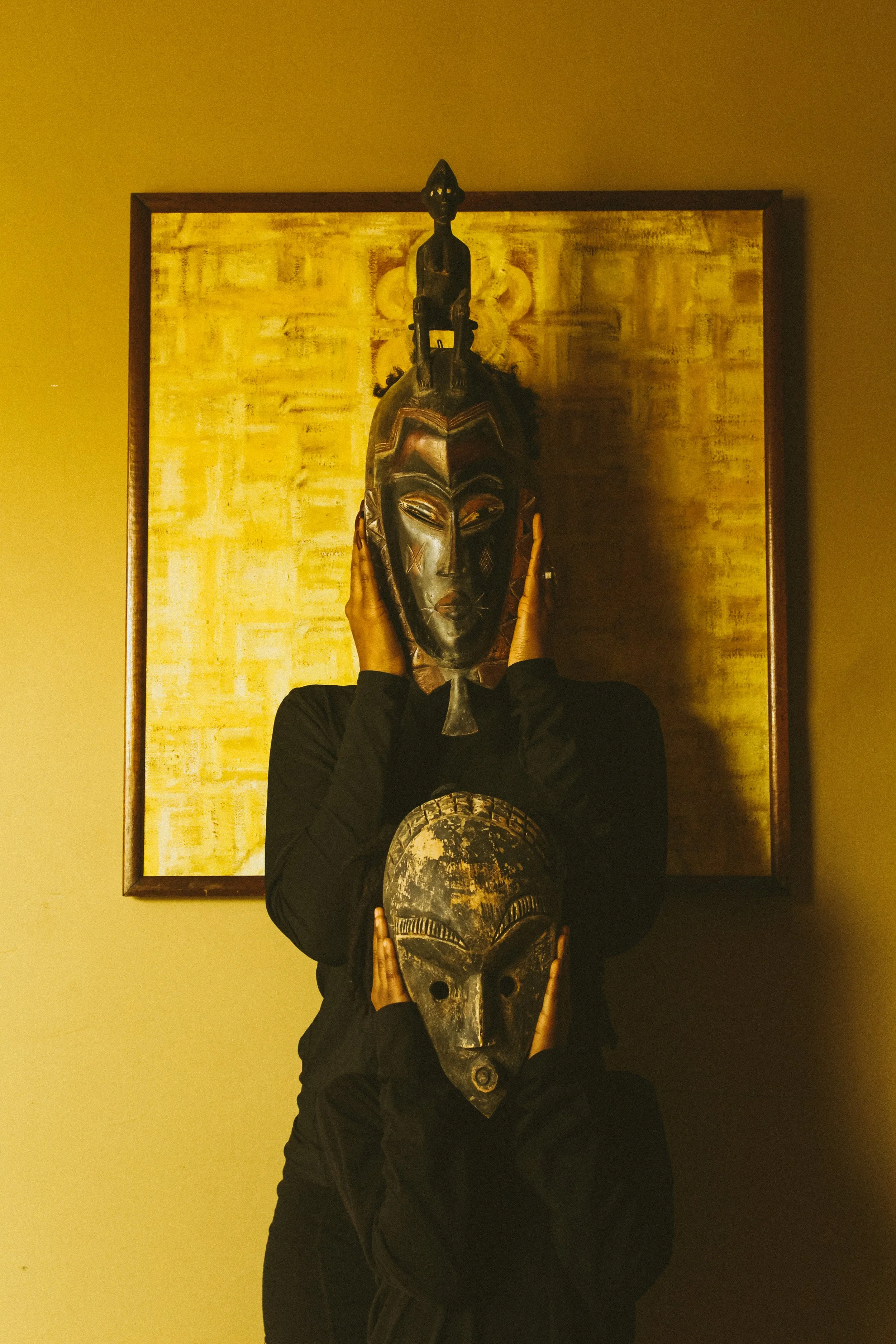 Person holding African tribal masks in front of a yellow and gold artwork.