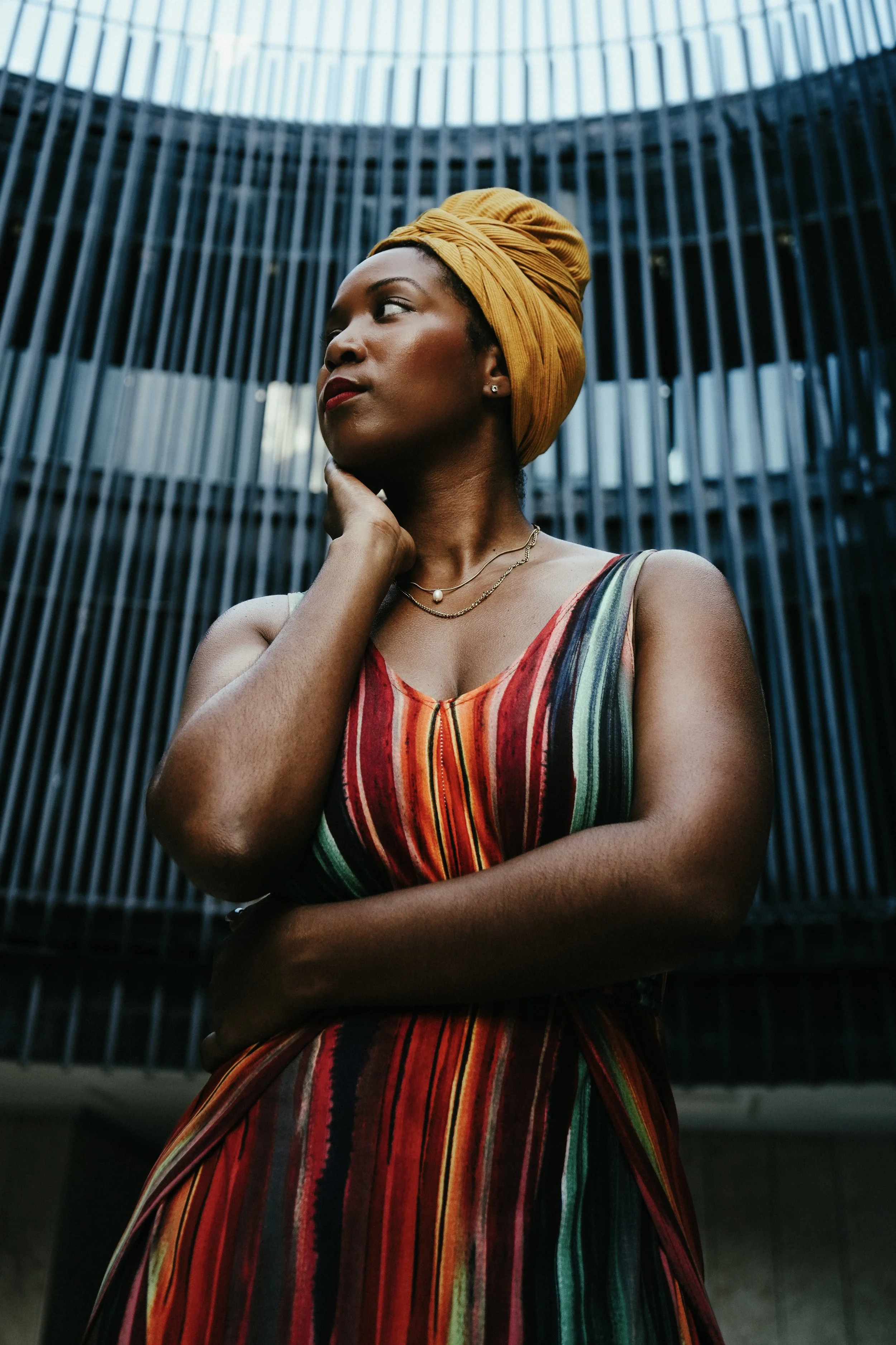 A woman with dark skin wearing a yellow headwrap and a colorful striped dress, standing outdoors in front of a modern architectural structure with vertical metal bars.