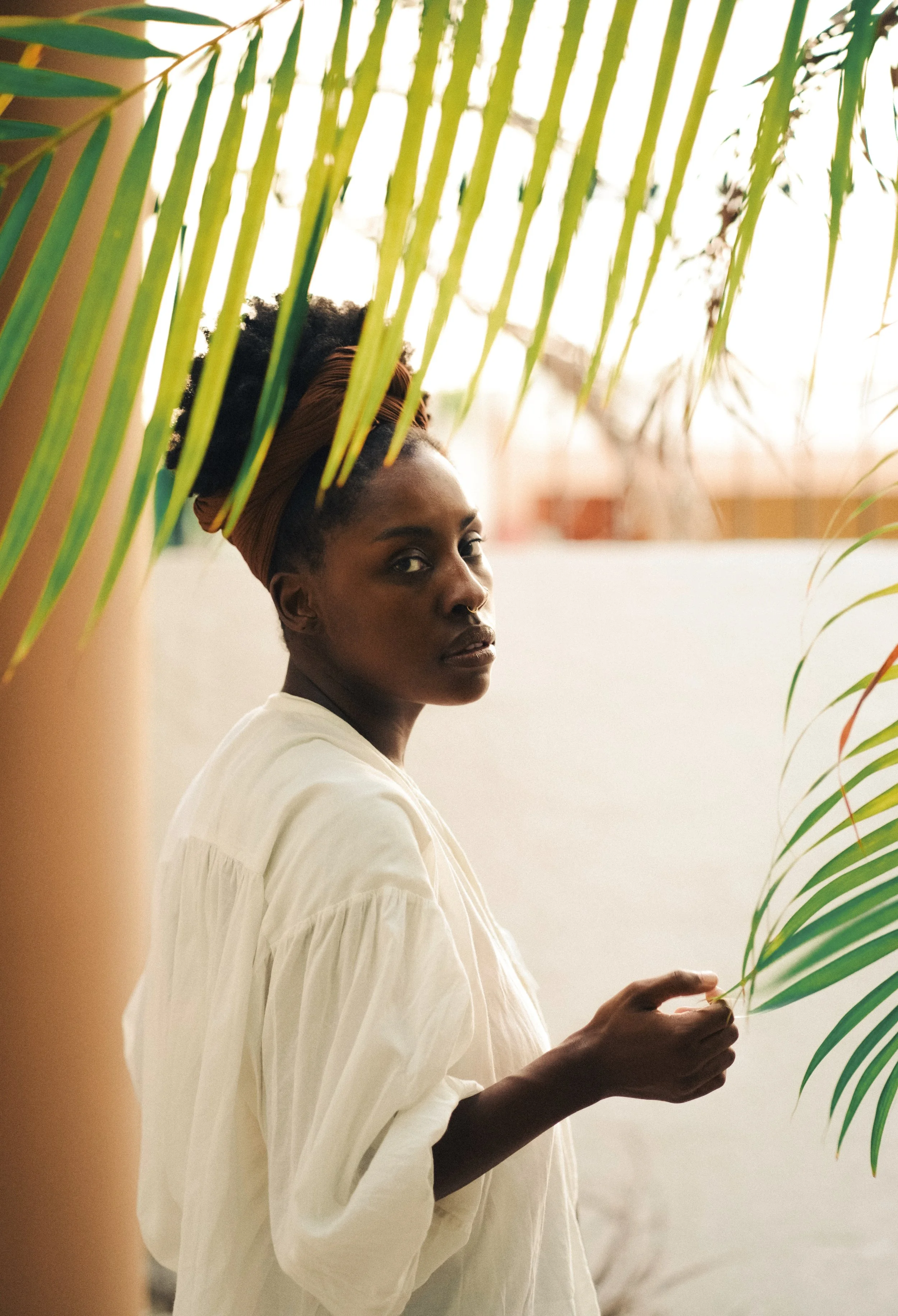 A woman with dark skin, wearing a white blouse and a brown headwrap, stands behind green palm leaves, holding a leaf in her right hand, in front of a light-colored wall.