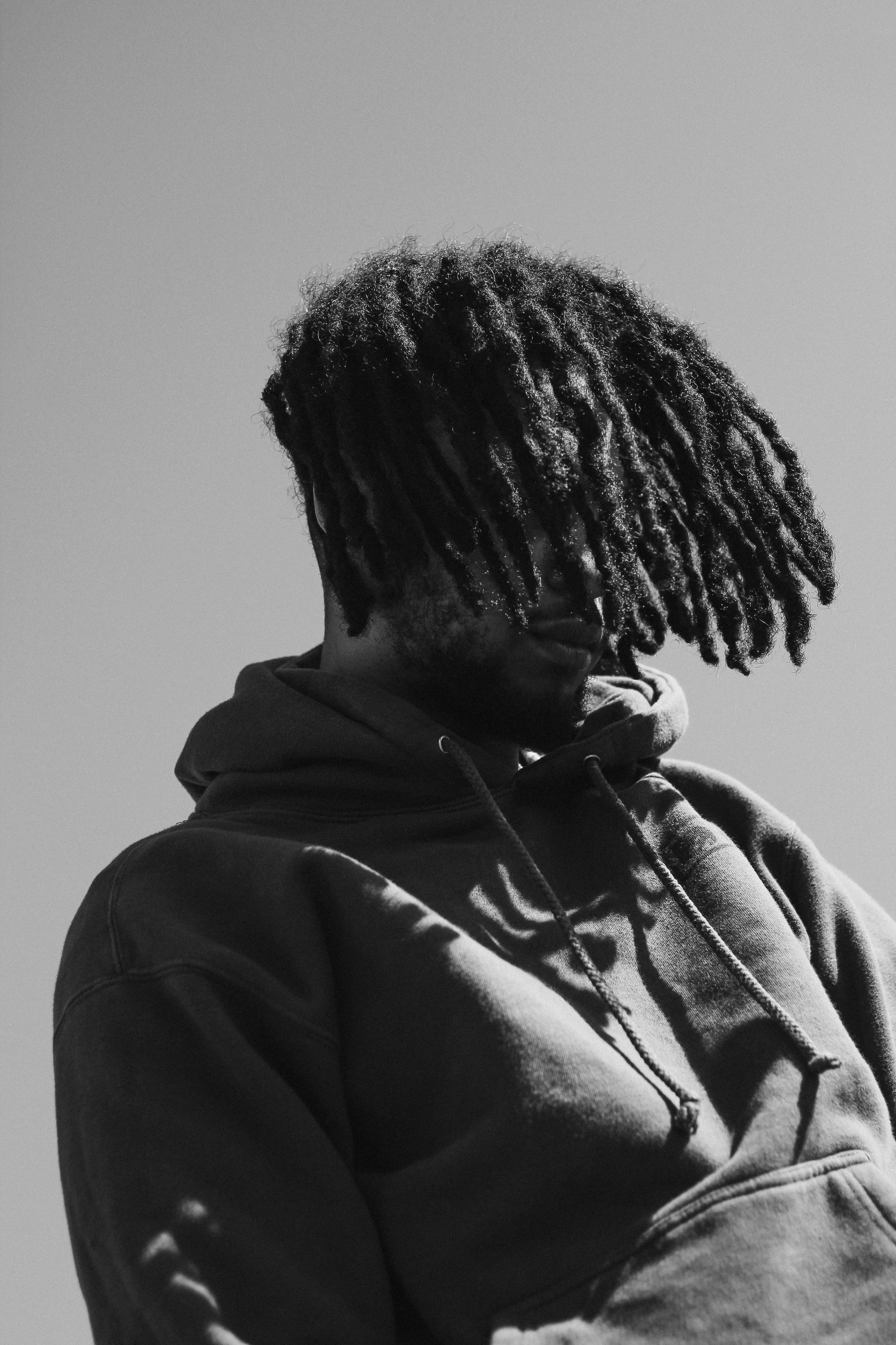 Black and white photo of a man with dreadlocks wearing a hoodie, facing slightly to the side, against a plain background.