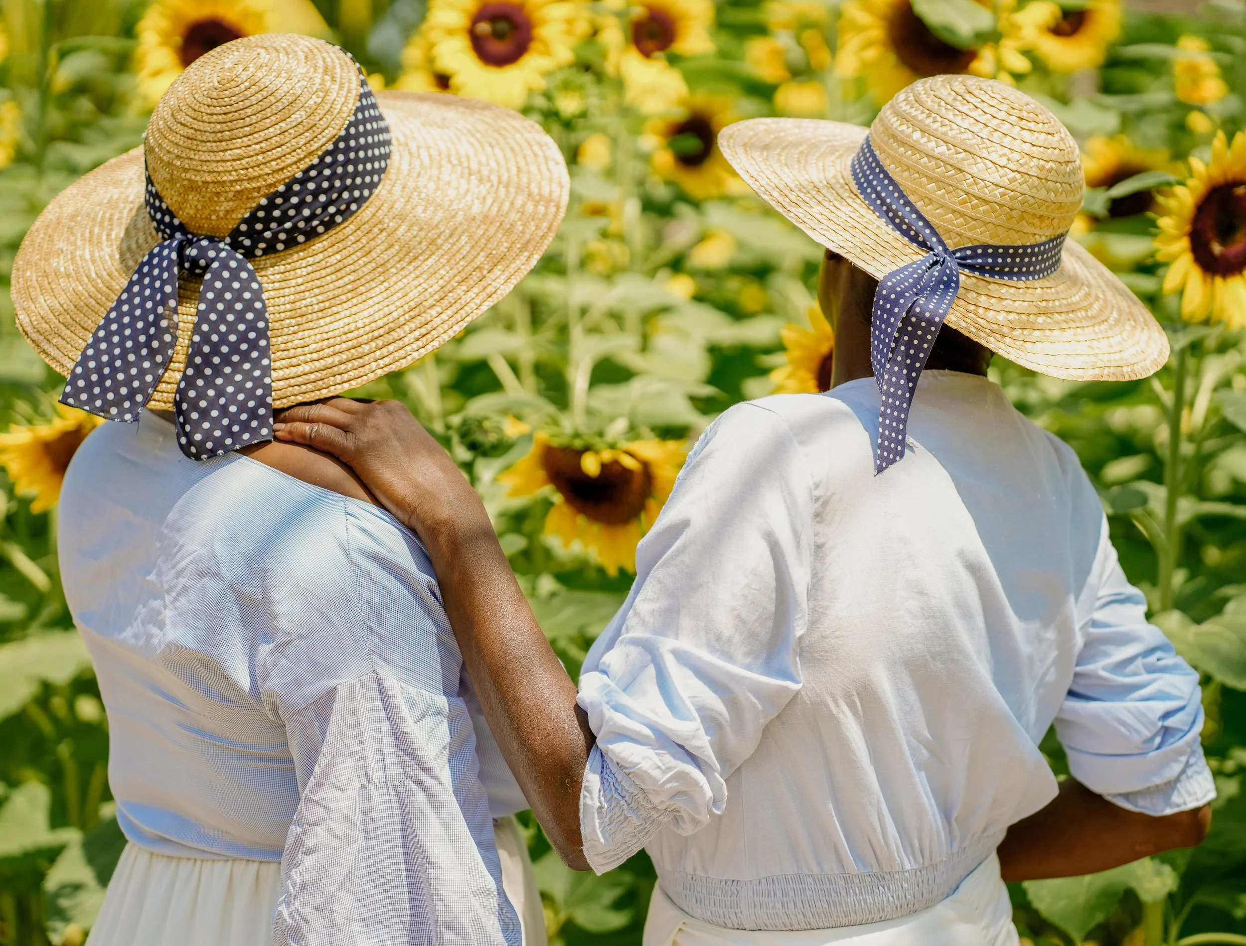 Two women wearing wide-brimmed sun hats with polka dot ribbons, standing close together in a sunflower field.