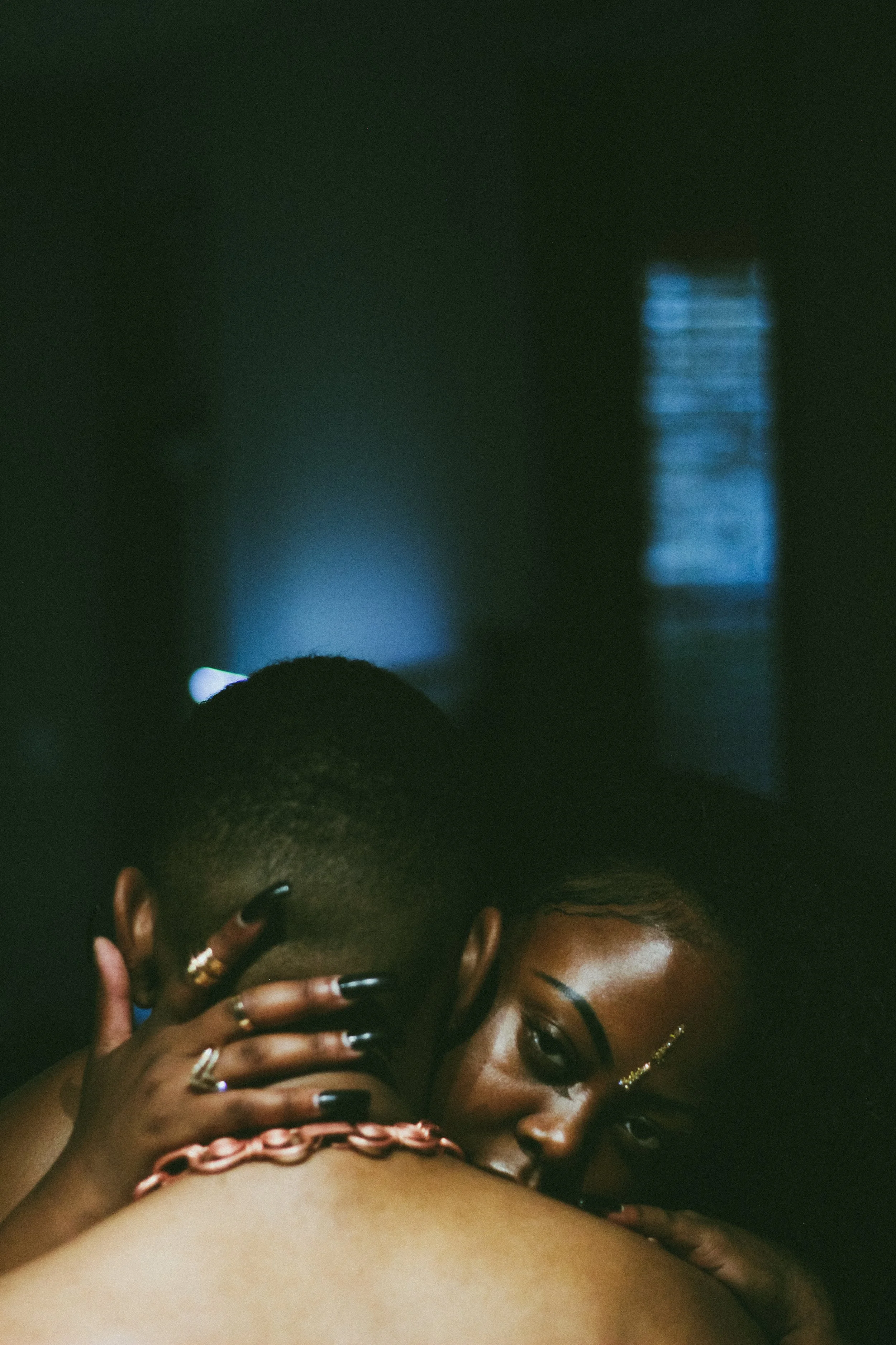 A close-up of a woman with dark skin and glasses, with her hand on her face, wearing jewelry, and a man with dark skin and short hair also close to her face, with a dark background and a blue light window in the distance.