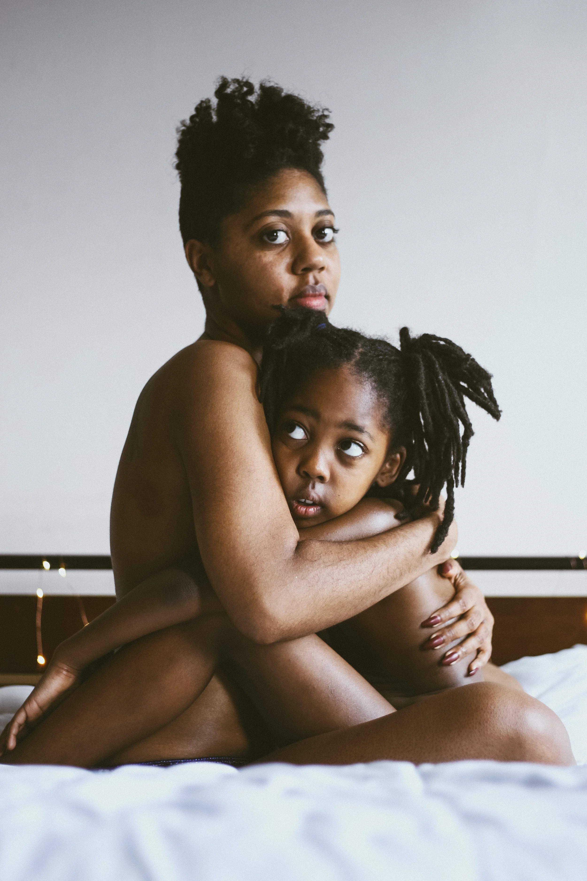 A woman and a child hugging each other on a bed with a headboard decorated with string lights.