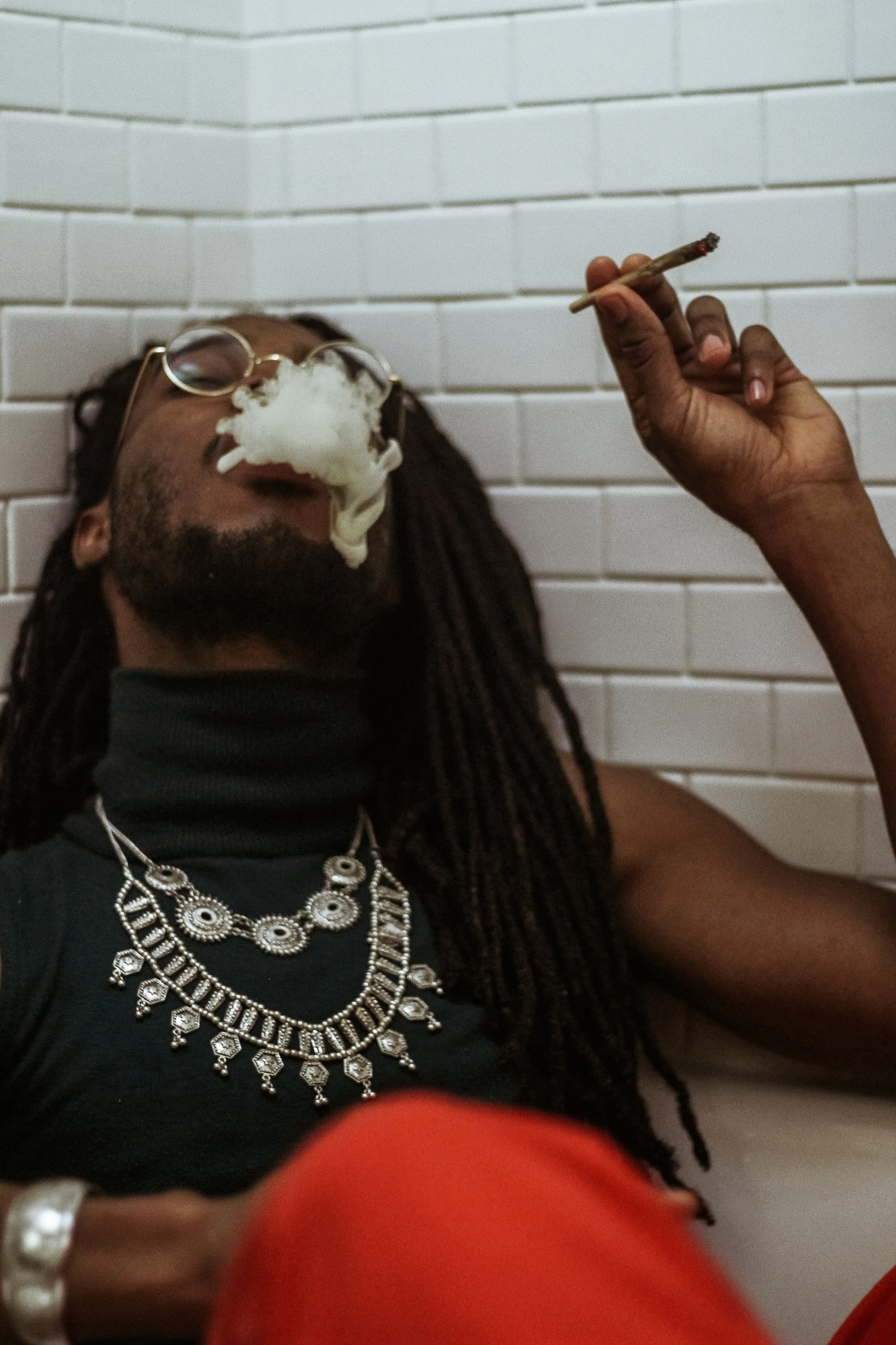 A person with braided hair and glasses is leaning back against a white tiled wall, smoking a joint with smoke billowing around their face. They are wearing jewelry, including multiple silver necklaces and rings, and a black shirt with red pants visib