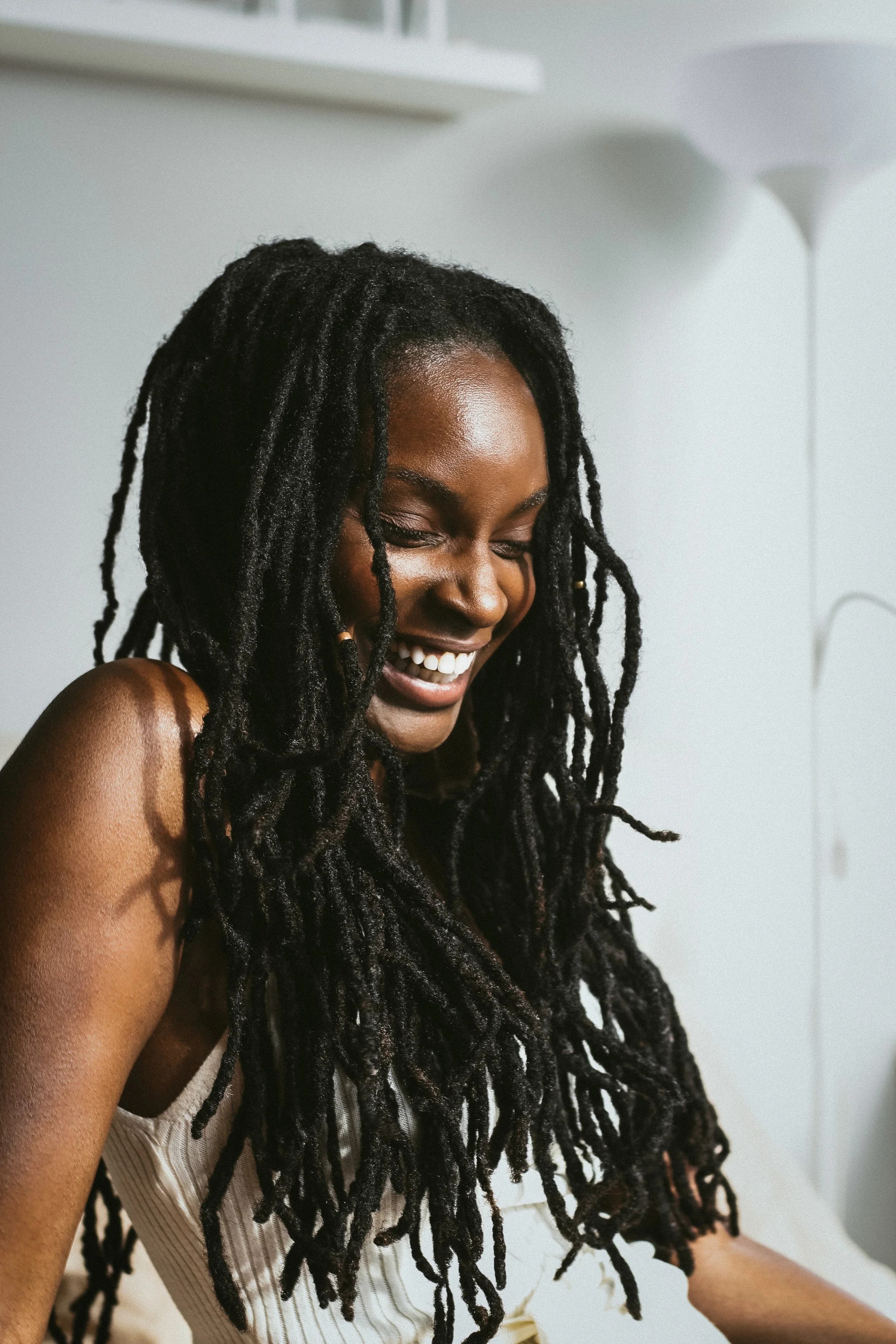 A woman with long dreadlocks is smiling with her eyes closed, wearing a sleeveless beige top, and standing indoors against a white wall with a white shelf and lamp in the background.