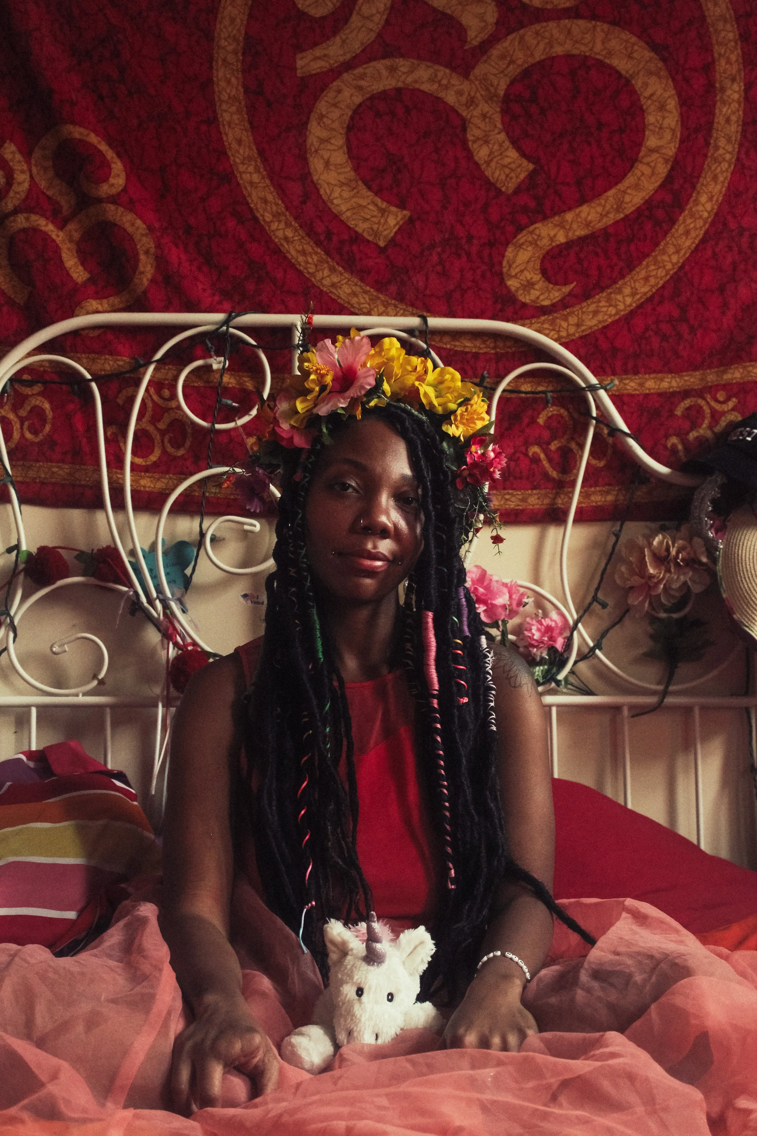A woman with long braided hair decorated with a flower crown, sitting on a bed with pink bedding, holding a plush unicorn toy, with a red wall hanging and fairy lights behind her.