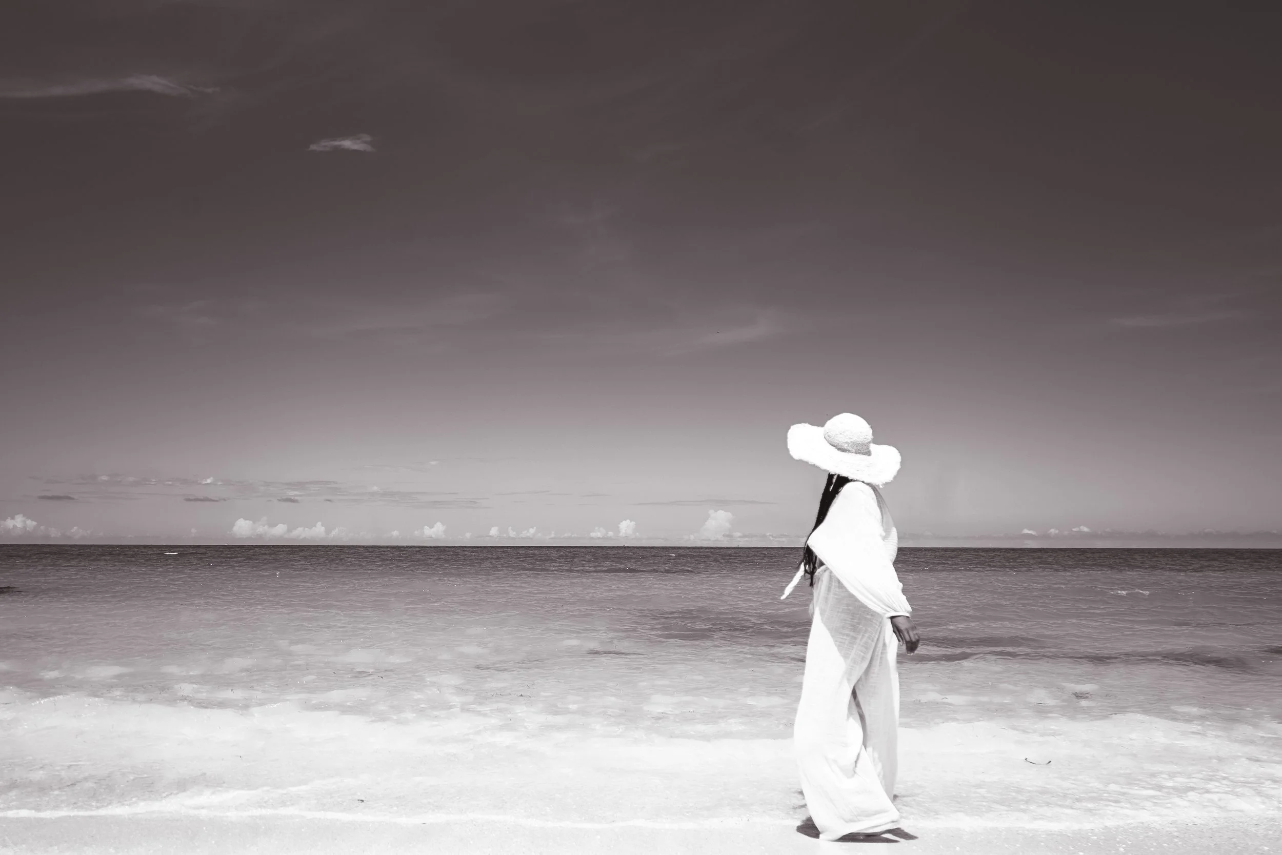 A woman wearing a large sunhat and light-colored clothing walking along the shoreline of a beach, with the ocean and sky in the background.