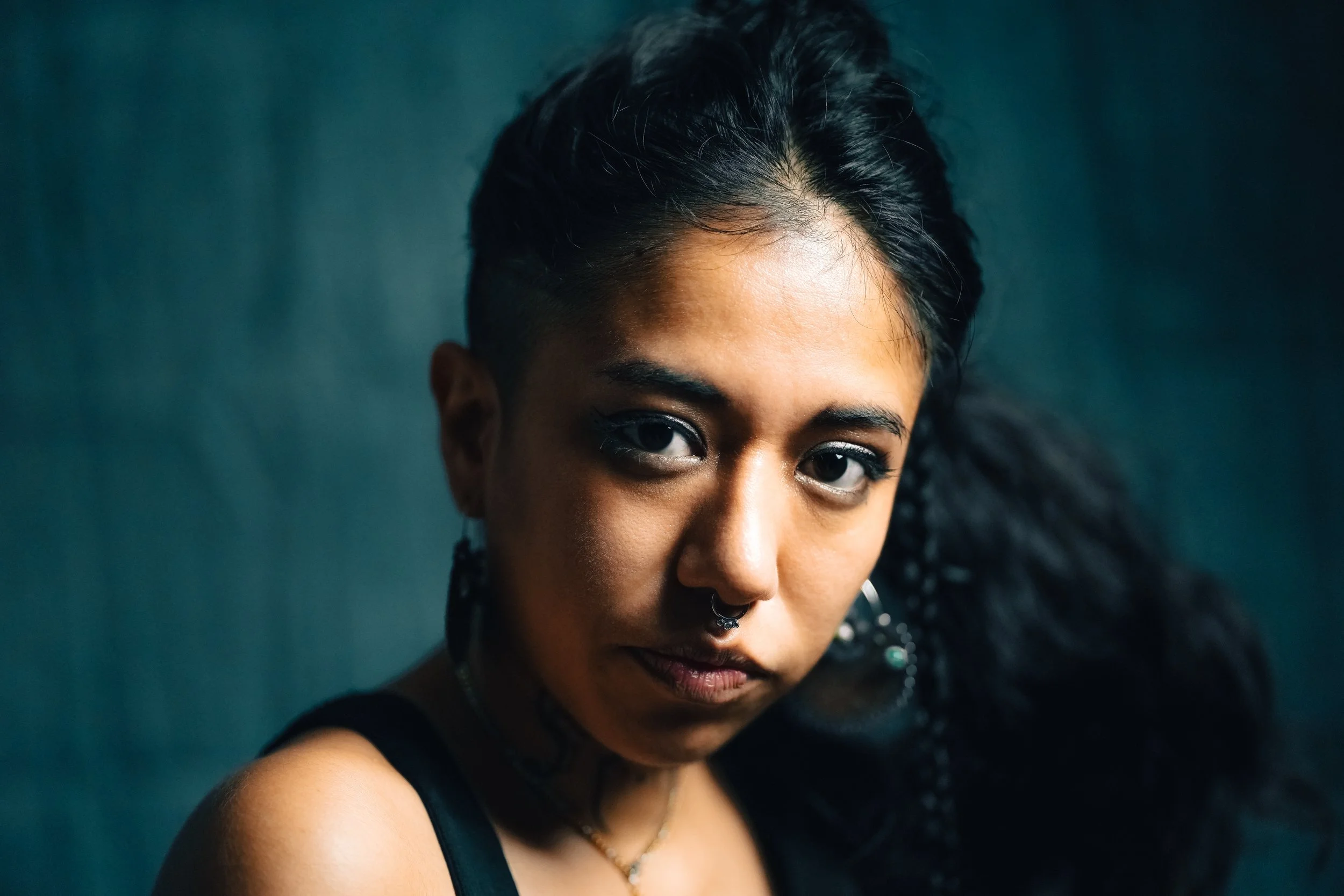 Close-up portrait of a woman with dark wavy hair, dark eyes, and a septum piercing, wearing hoop earrings and a black top, against a dark background.