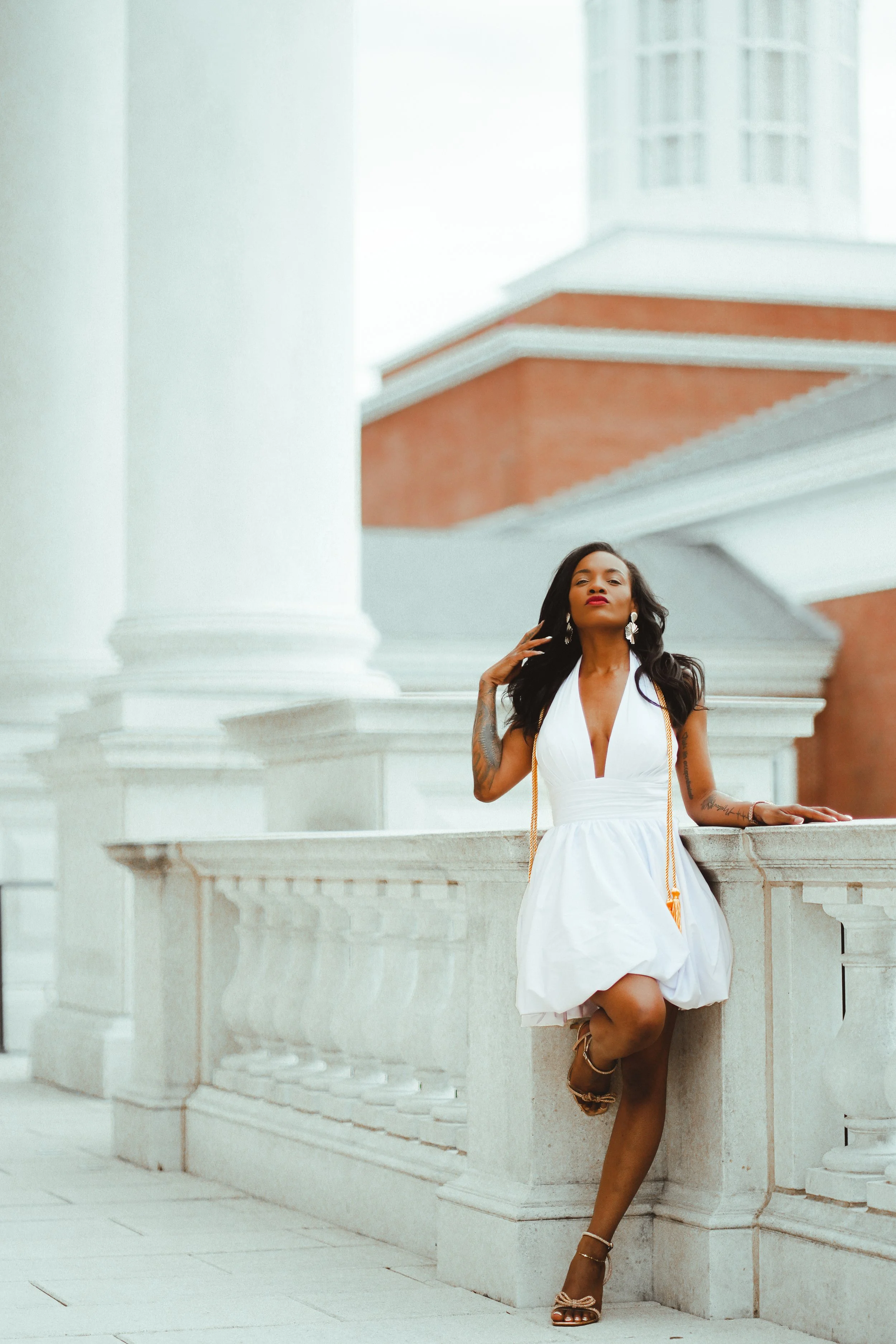 A woman in a white dress posing on a stone architectural structure with columns and stairs, with a large building with windows in the background.