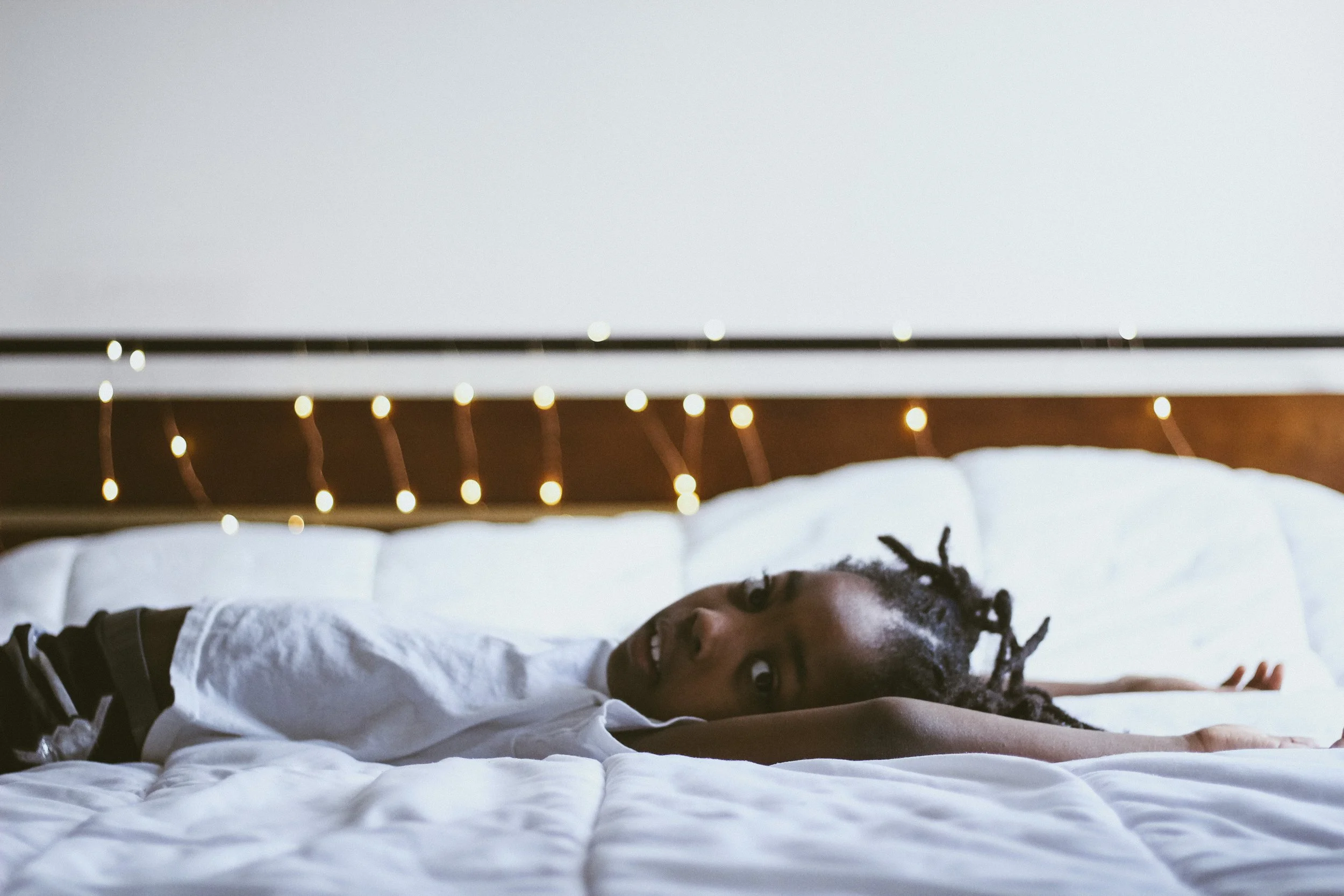A woman with braided hair lying on a bed, looking to the side. The bed has white bedding, and the background features a headboard with string lights.