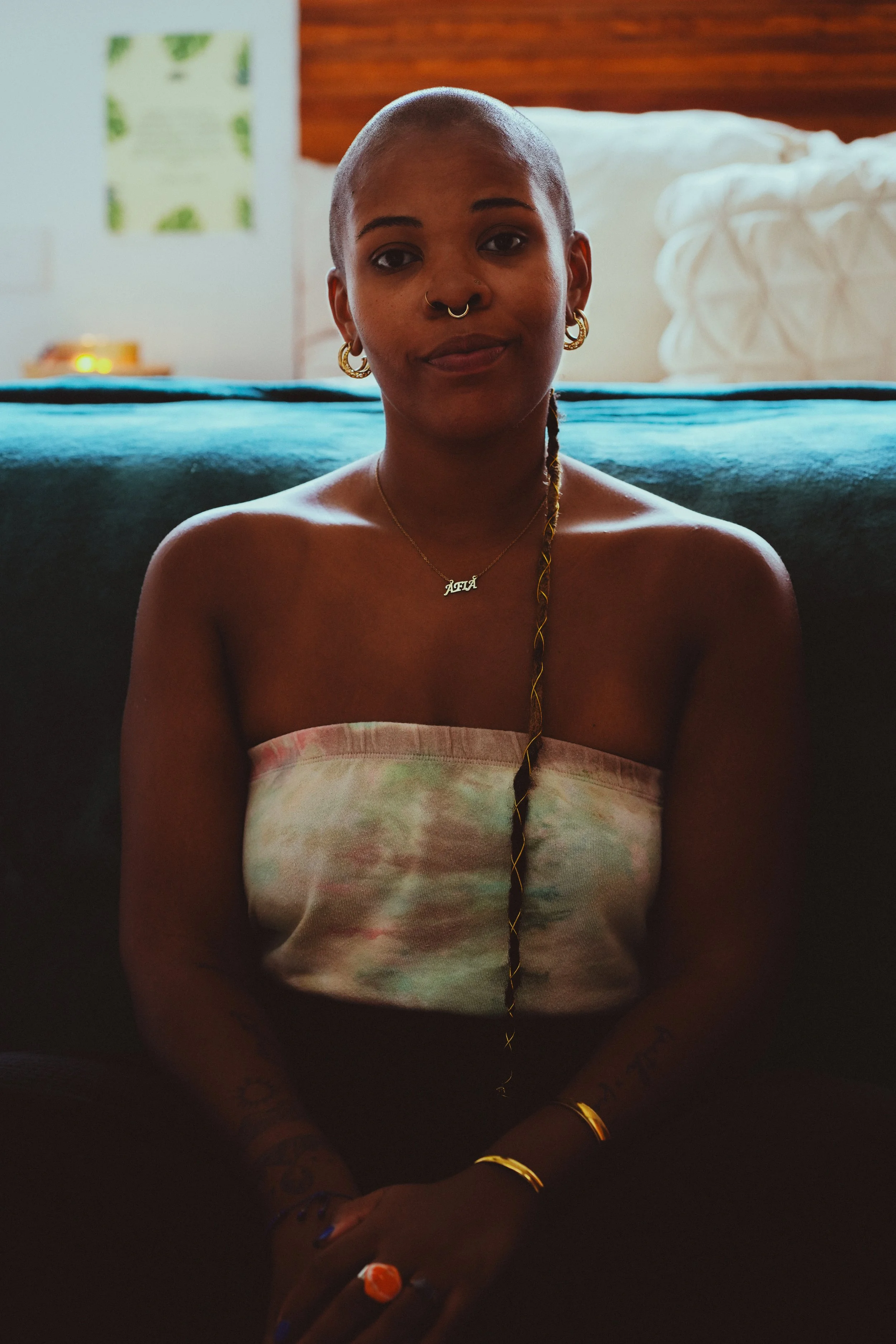 A woman with a shaved head and multiple gold jewelry, including earrings, necklace, and bracelets, sitting in a dimly lit room with pillows and a bed in the background.