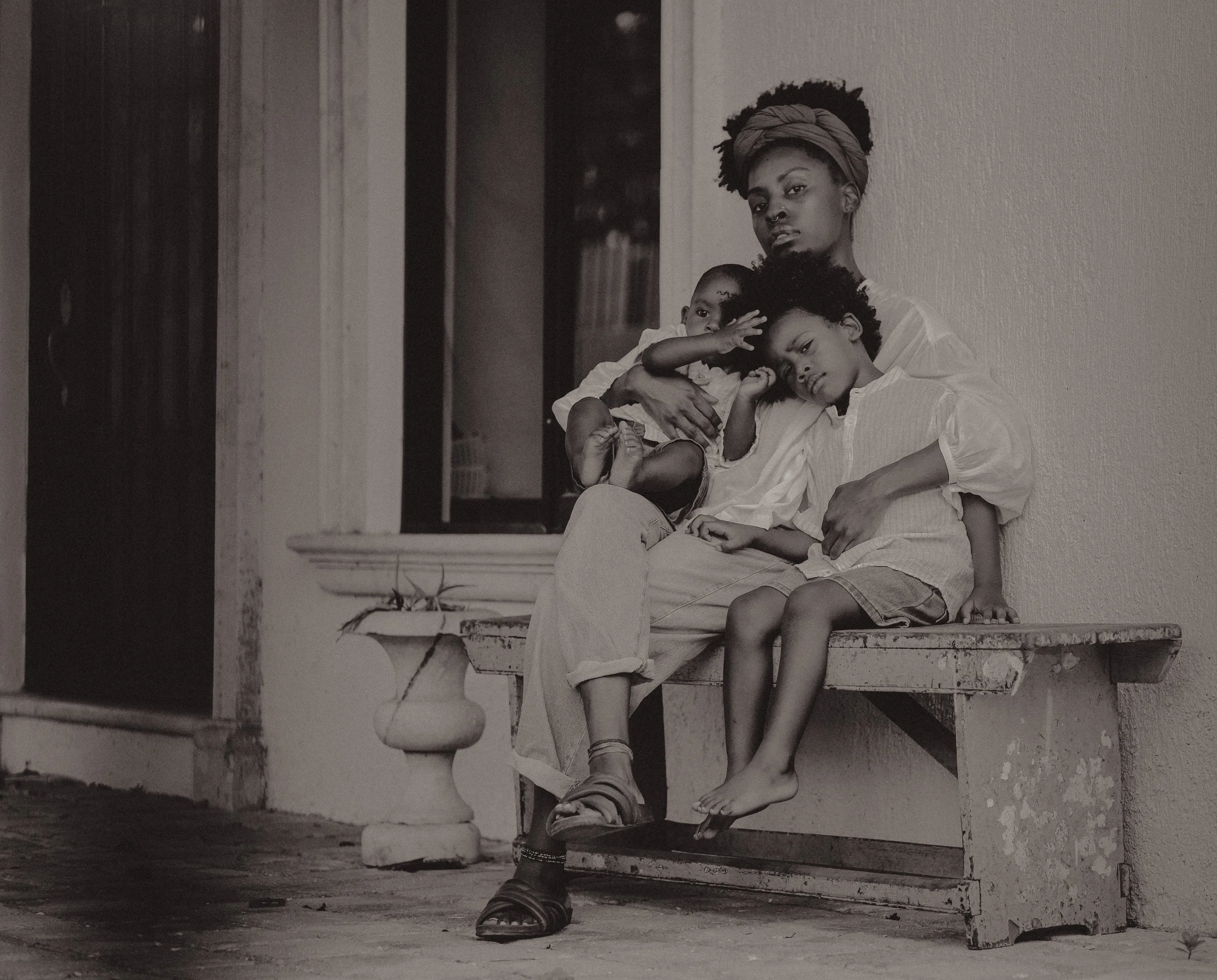Black and white photo of a woman sitting on a bench outside, holding two children on her lap. The woman has curled hair wrapped in a headscarf. One child is resting on her shoulder, and the other is sitting beside her, leaning head on her arm. The ch
