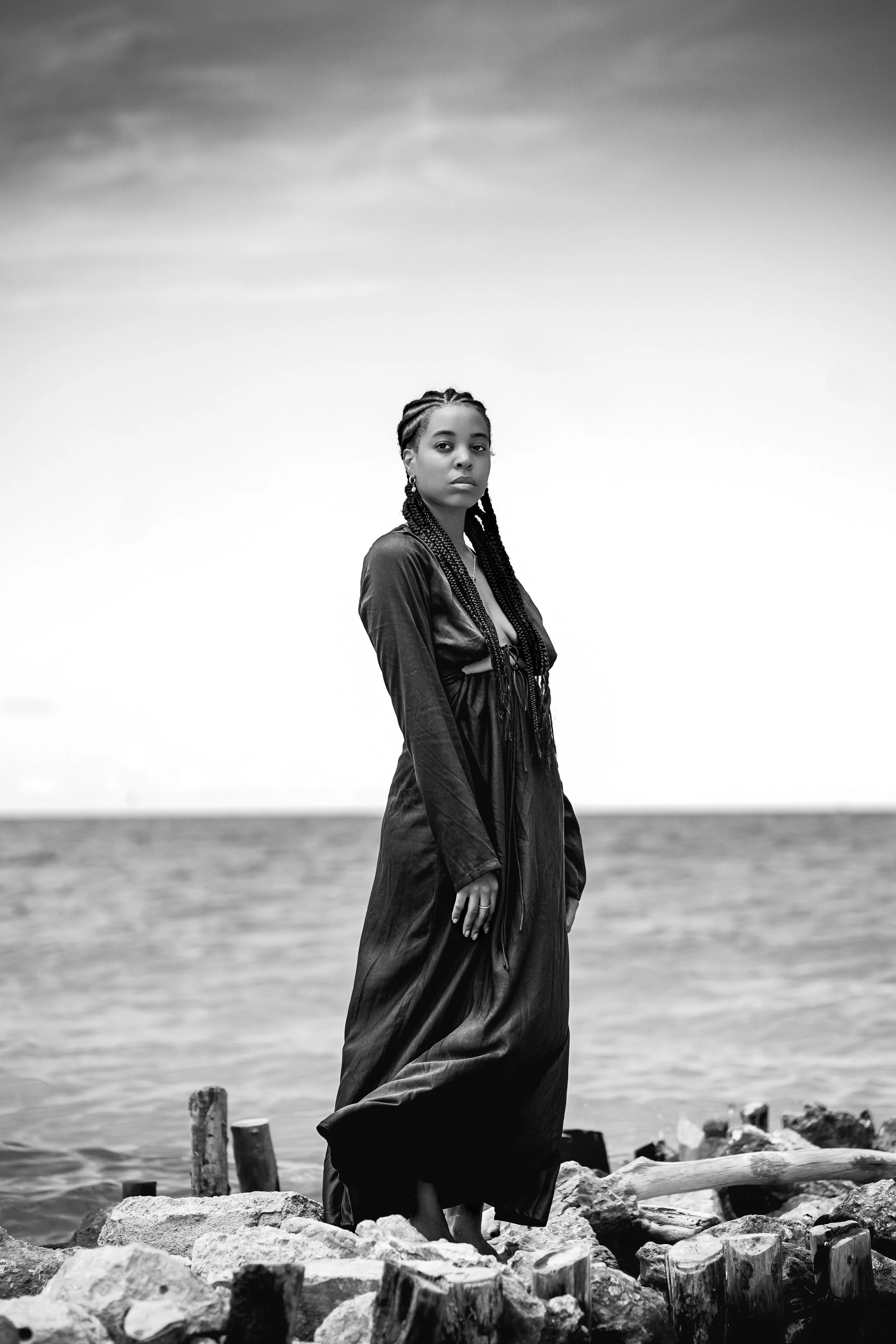 A woman with braided hair and earrings standing on rocks by the water, wearing a long dress or coat, looking at the camera with a serious expression, in black and white.