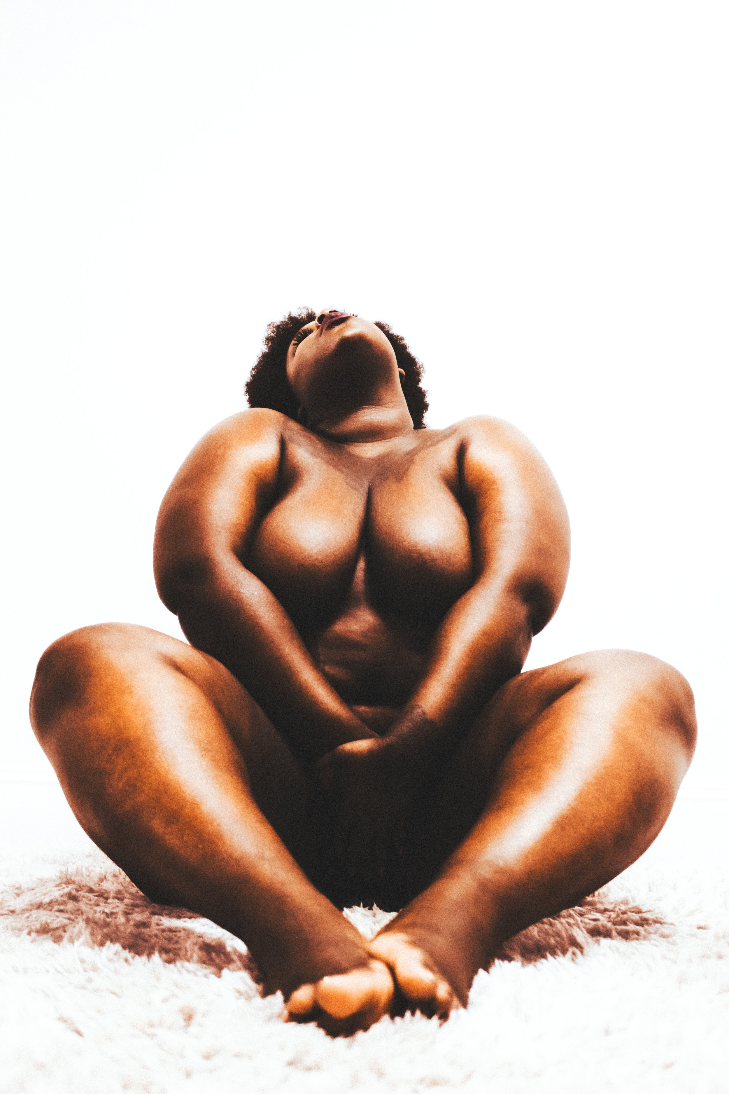 A woman with dark skin and curly hair sitting cross-legged on a soft rug, looking upwards with her eyes closed and hands resting on her lap, against a plain white background.