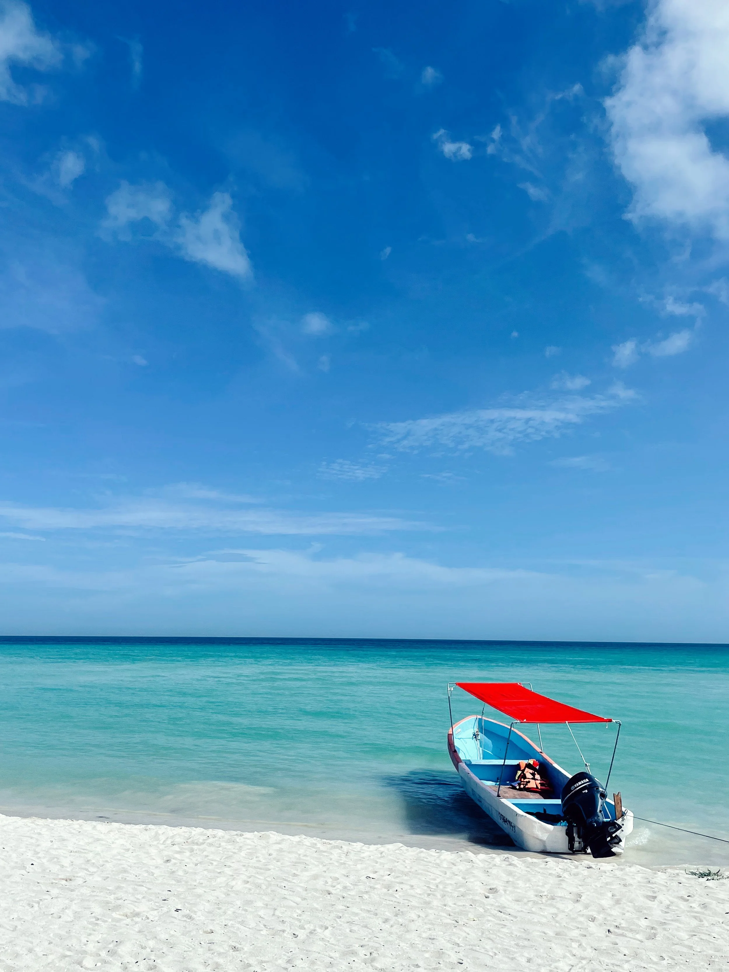 A boat with a red canopy resting on the white sandy beach, turquoise water, and a blue sky with some clouds.