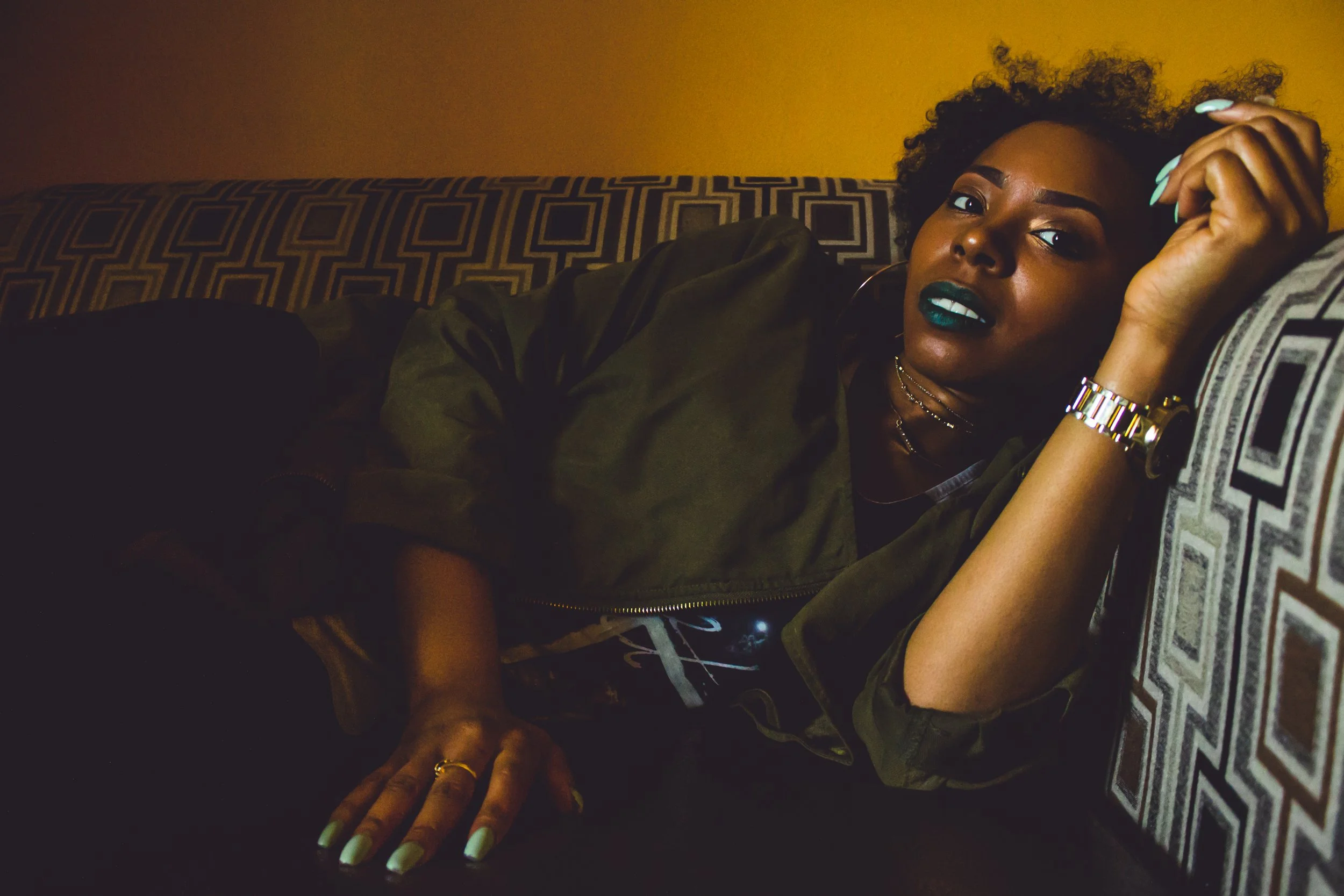 A woman with dark curly hair, dark lipstick, and gold jewelry lying on a patterned couch in warm lighting.