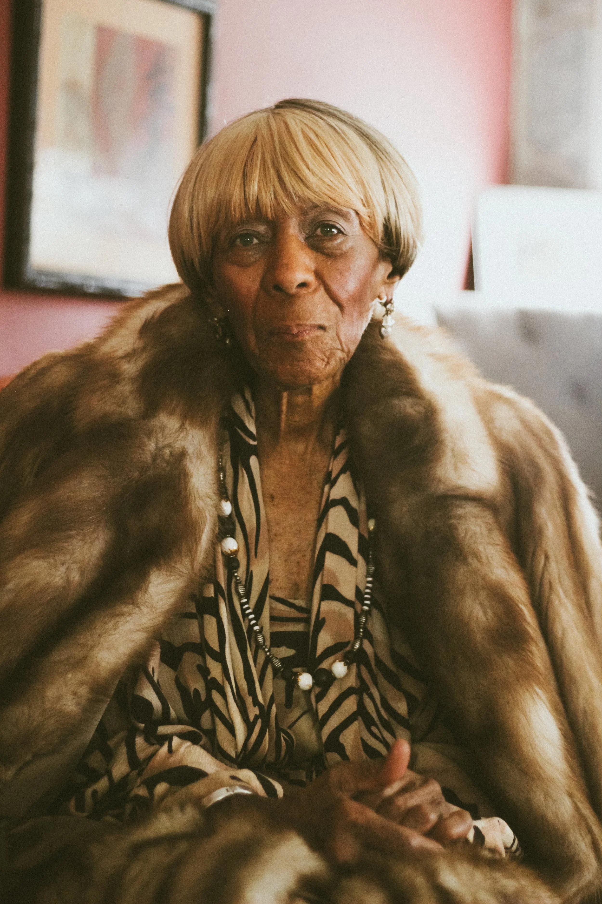 A woman with short blond hair and earrings wearing a fur coat and patterned blouse, sitting indoors.