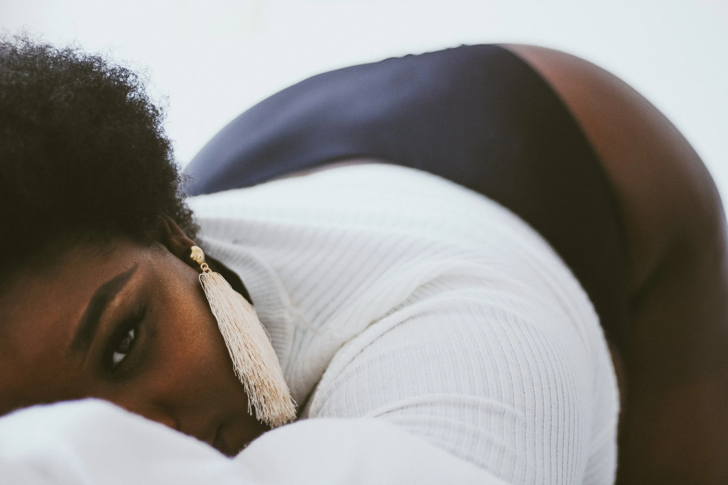 Close-up of a person with dark skin, black curly hair, wearing a white long-sleeve top, and a large white tassel earring, lying on a white surface.
