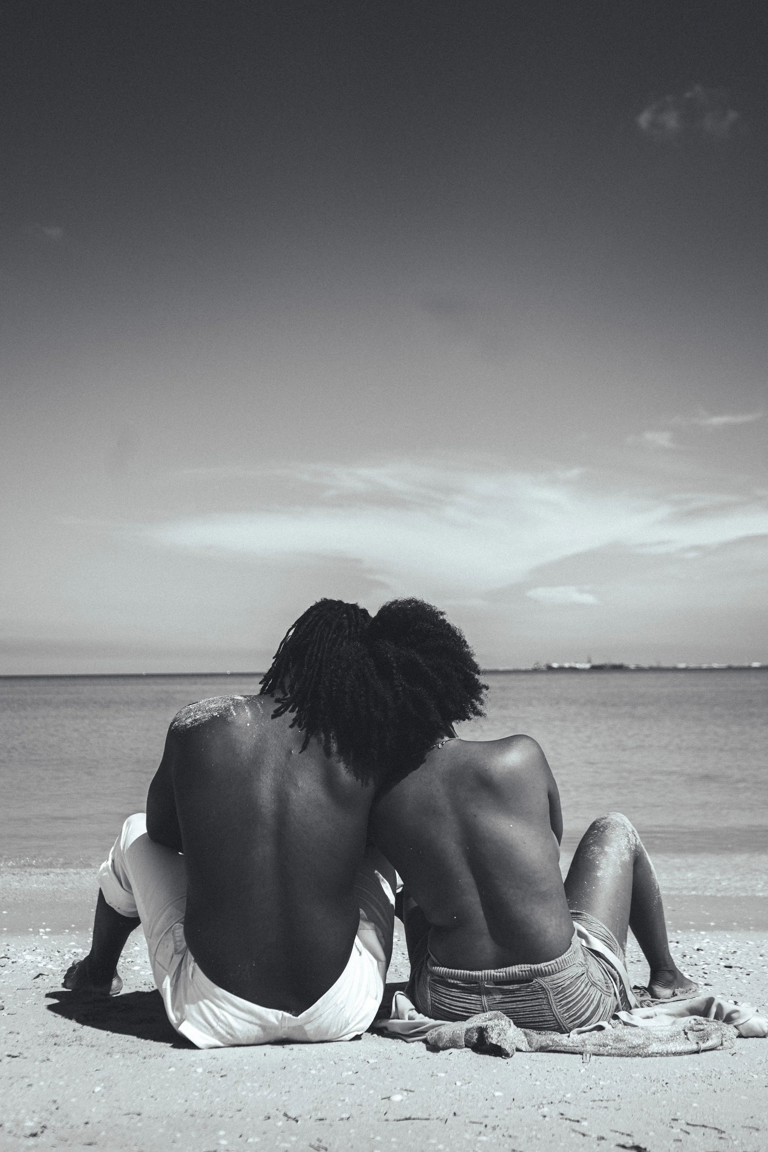 A black and white photo of two people sitting on a beach, facing the ocean, with their arms around each other. One person is shirtless with dreadlocks, wearing light-colored shorts, and the other person is wearing striped shorts.