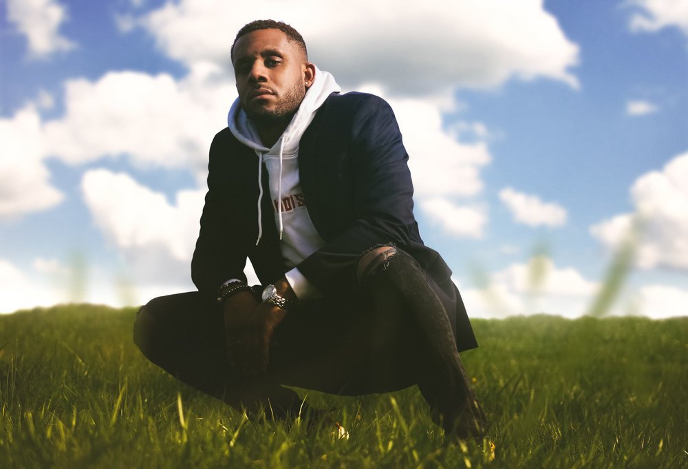 Young man crouching on grass field under blue sky with scattered clouds, wearing a hoodie, jacket, and jeans, looking at the camera.
