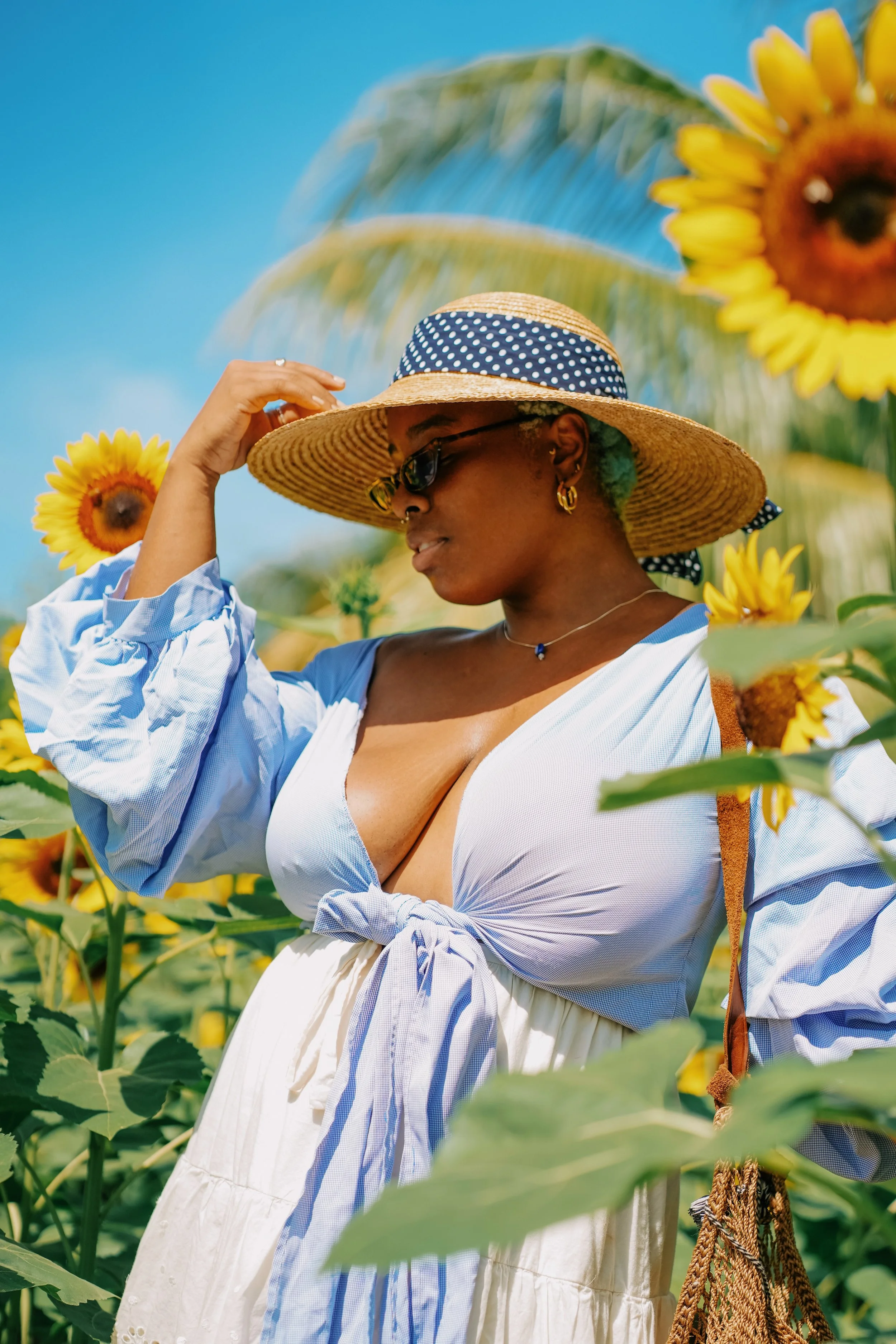 A woman wearing sunglasses, a wide-brimmed straw hat, and a blue and white outfit, standing among sunflowers and palm trees outdoors on a sunny day.