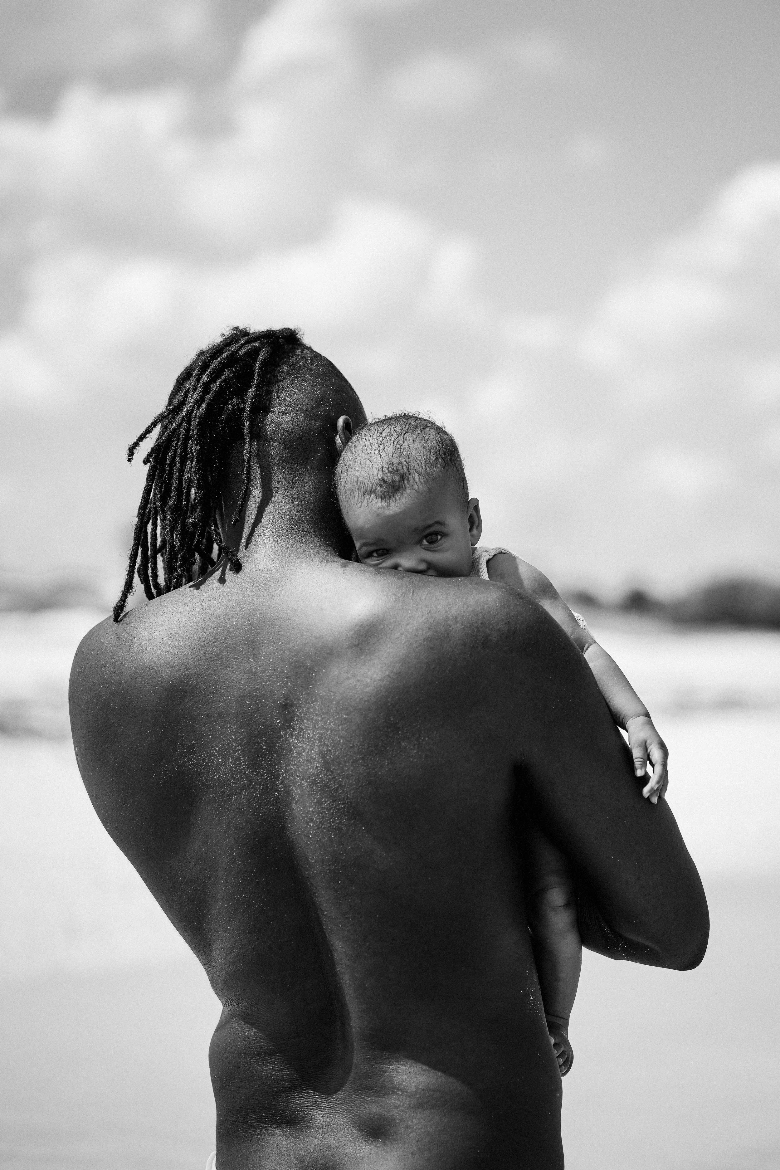 A person with dreadlocks holding a baby close to their shoulder at the beach, black and white photo, cloudy sky in the background.