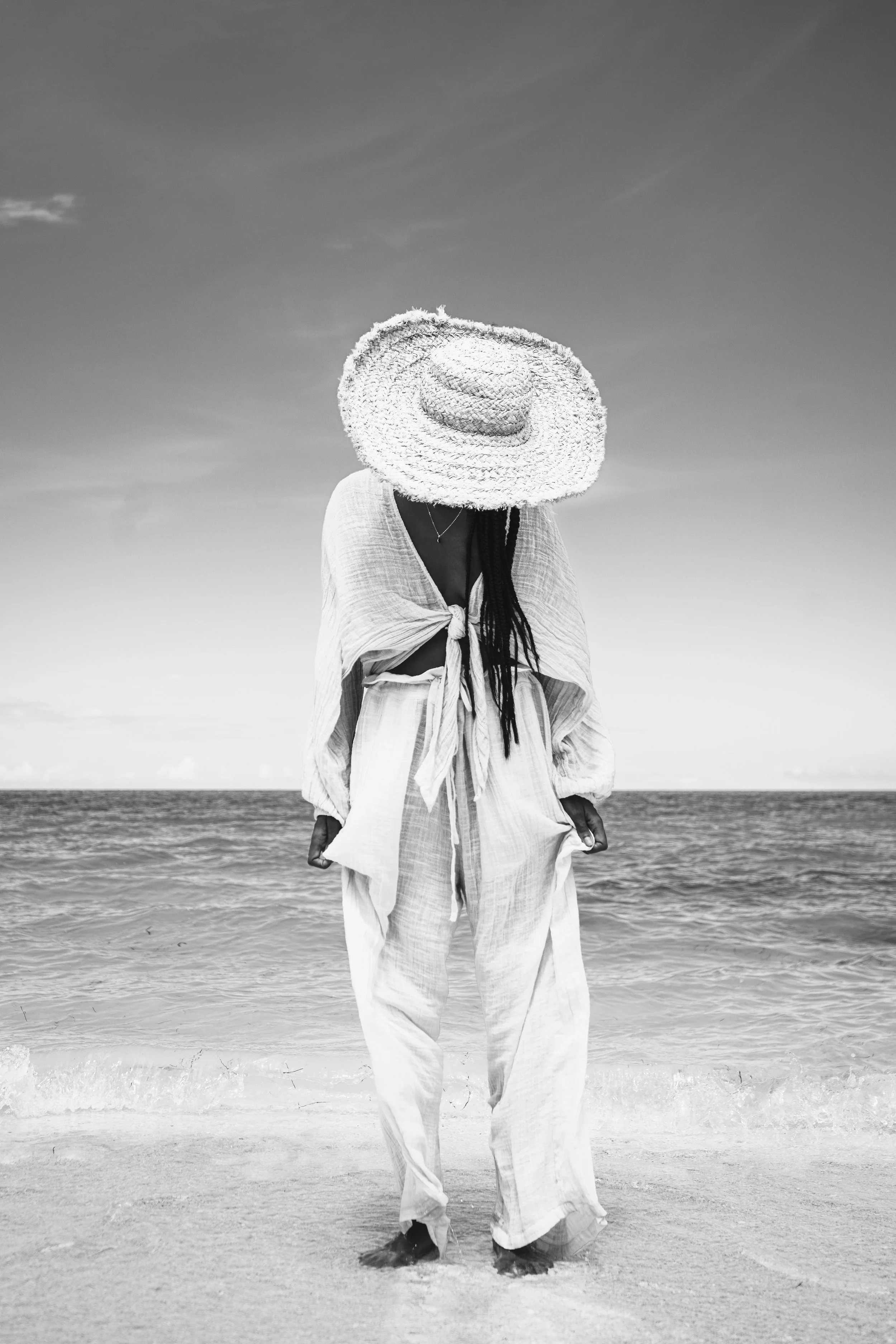 A person standing on the beach with a large straw hat covering their face, wearing loose, light-colored clothing, with the ocean and sky in the background.