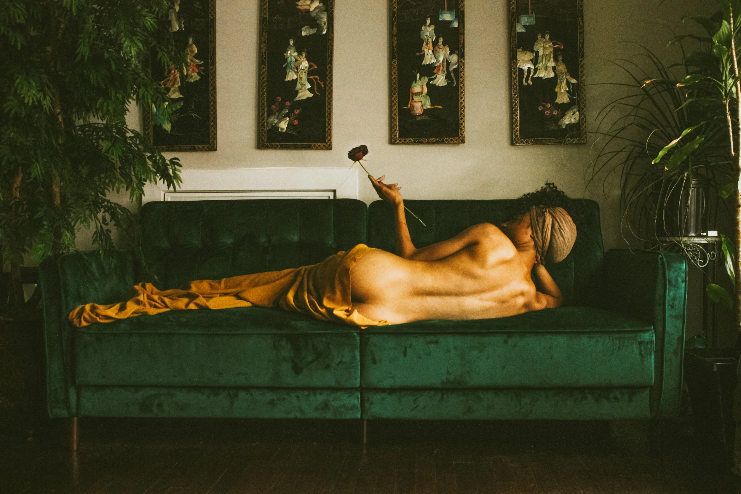 A reclining nude woman with a head wrap holding a single flower, lying on a green velvet sofa in a dimly lit room. Behind her is a wall decorated with five vertical panels of Asian art depicting figures in traditional attire. There are large green pl
