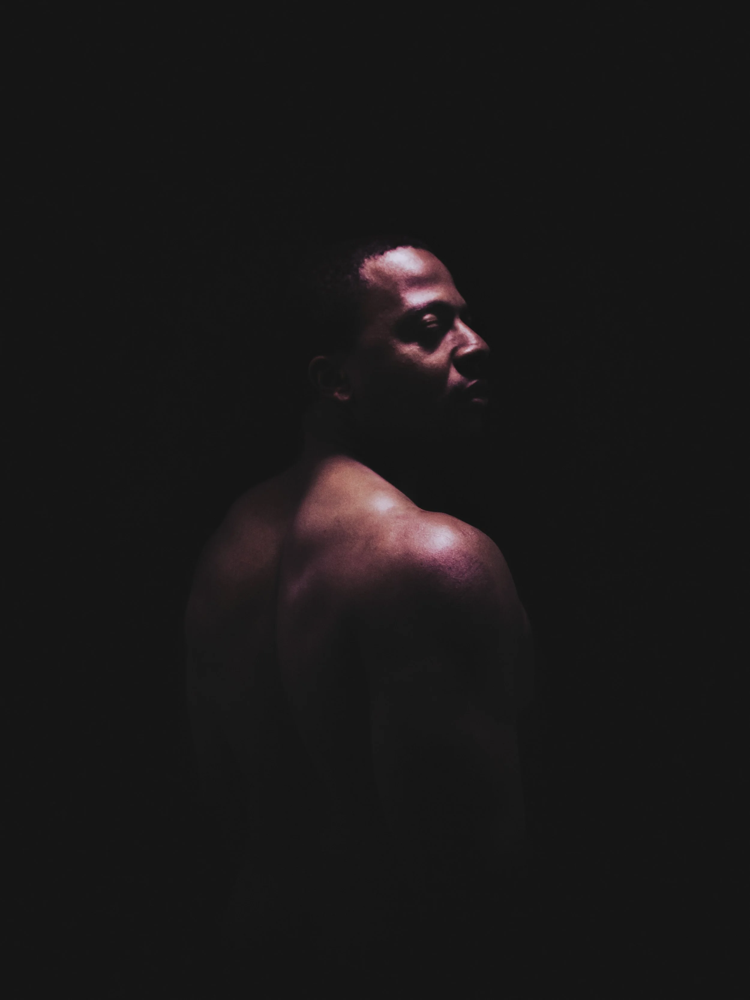 A woman with dark skin and minimalist makeup with closed eyes, photographed in side profile with dramatic lighting highlighting her face and shoulder against a black background.