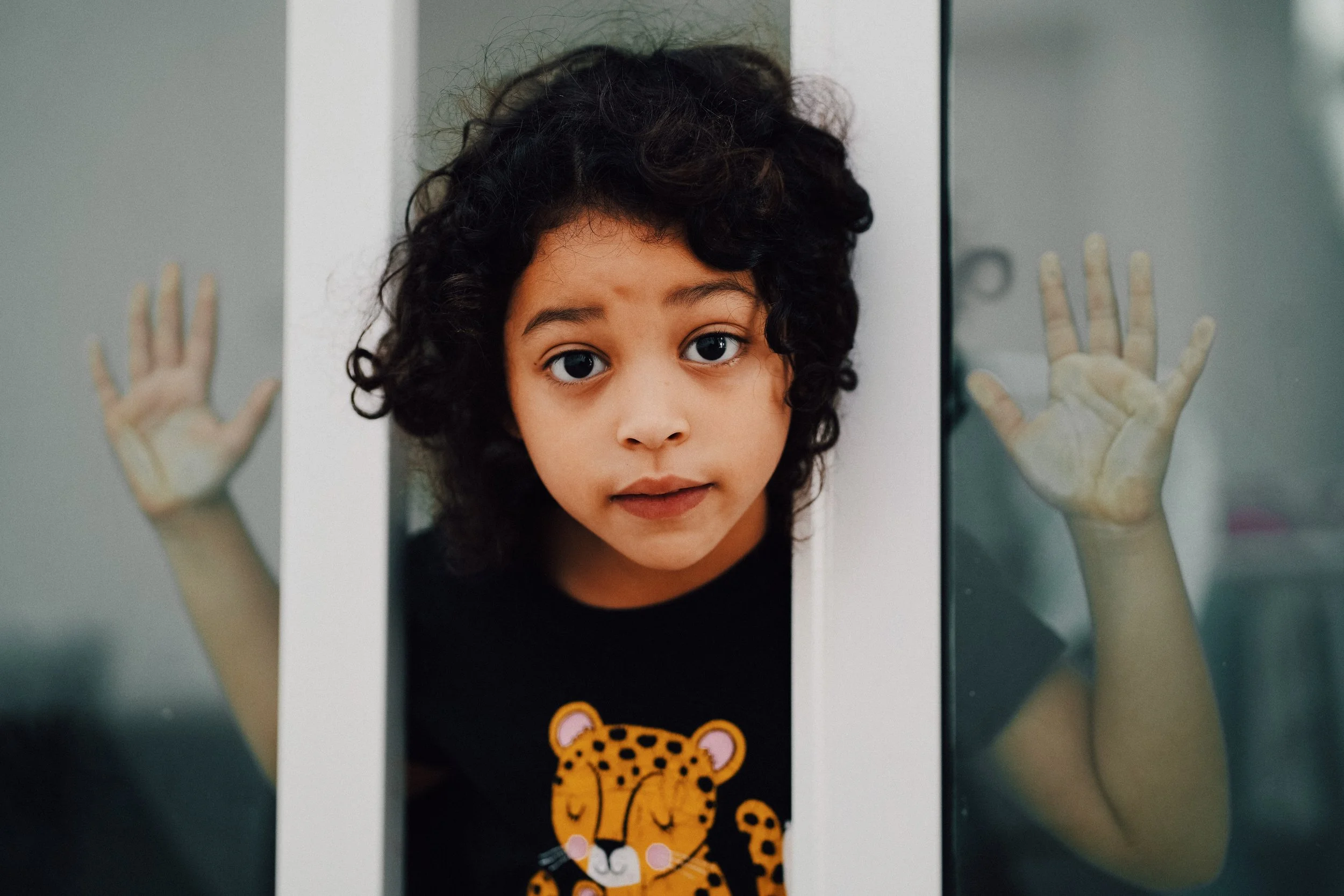 A young girl with curly dark hair and big eyes looking through an elevator door with her hands pressed against the glass.
