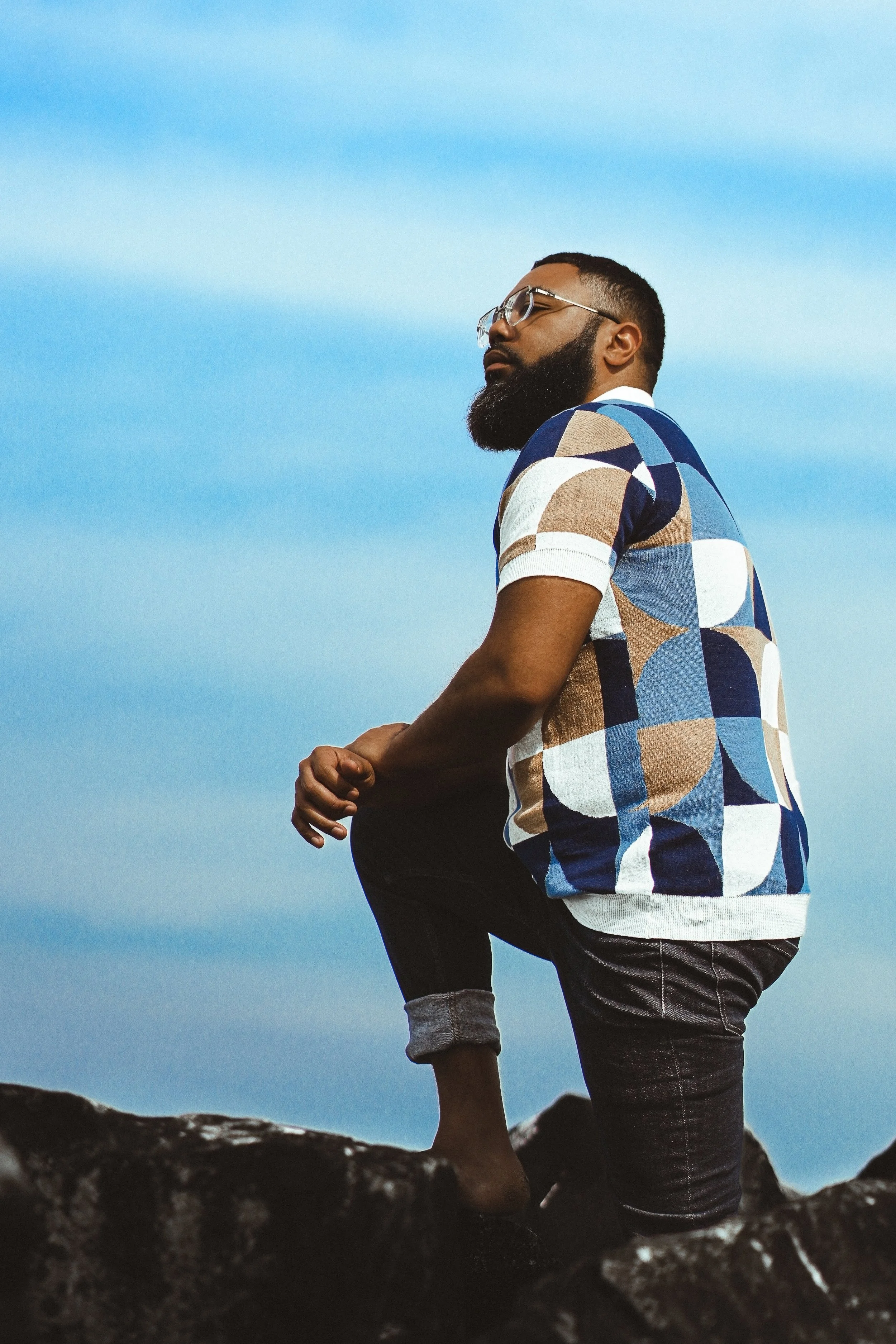 A man with glasses and a beard, wearing a colorful geometric patterned shirt, is sitting on a rock, with a clear blue sky in the background.