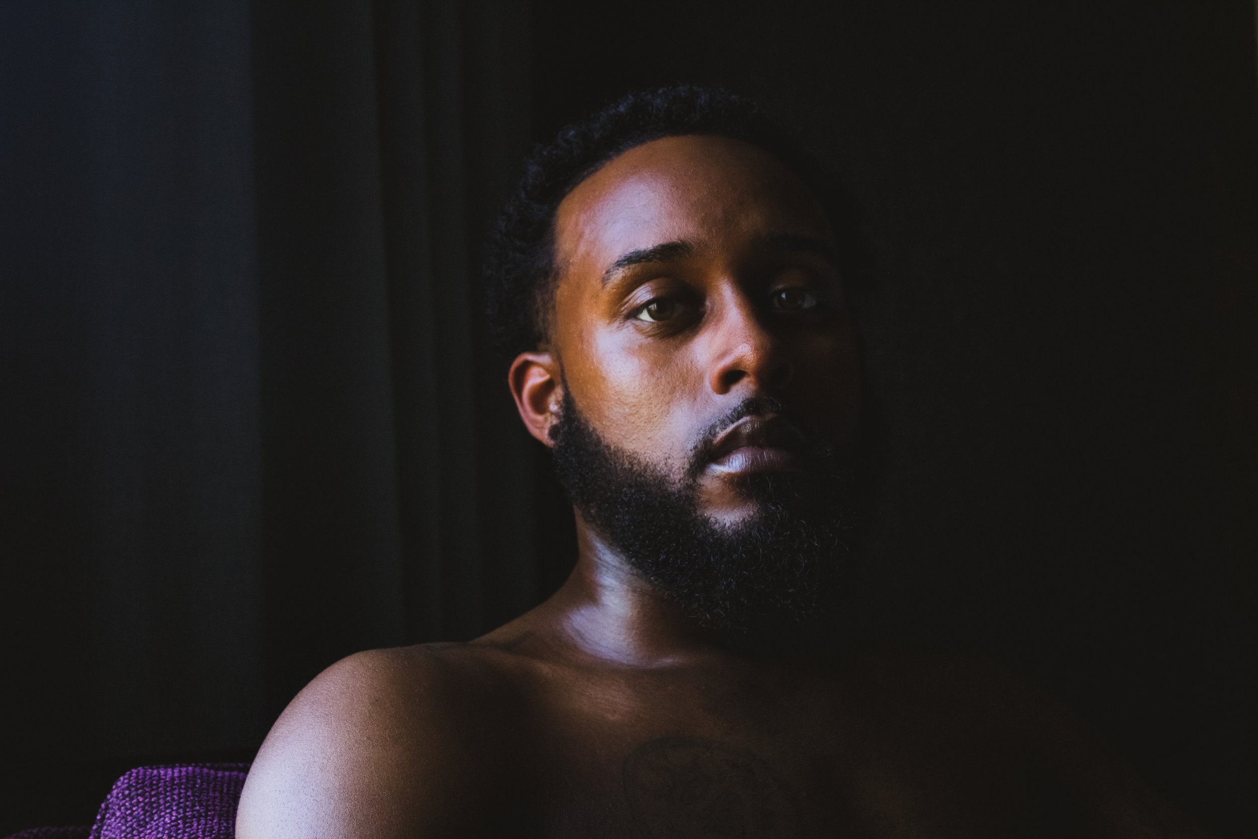Close-up portrait of a shirtless Black man with a beard and short curly hair, partially illuminated against a dark background.