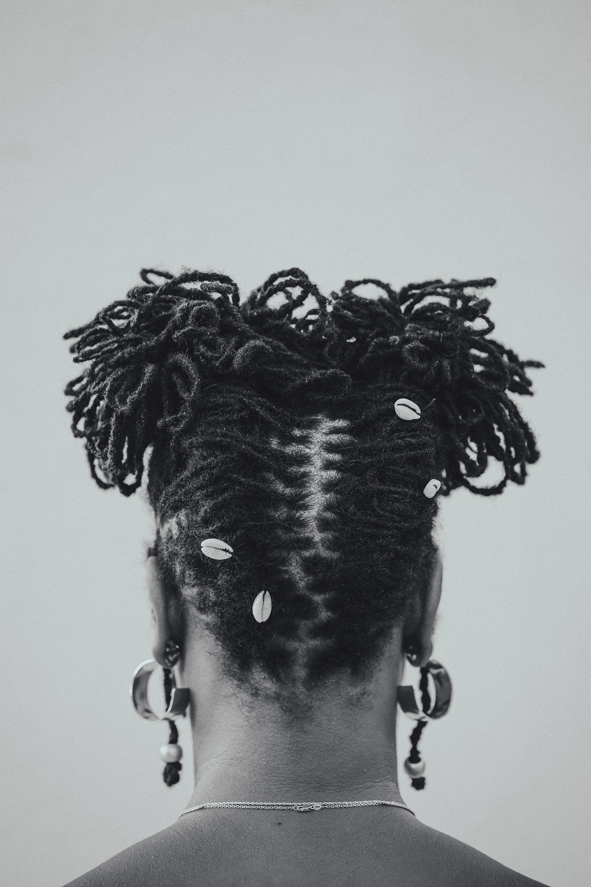 Back view of a person with styled dreadlocks adorned with beads and shells, wearing earrings and a necklace, against a plain background.