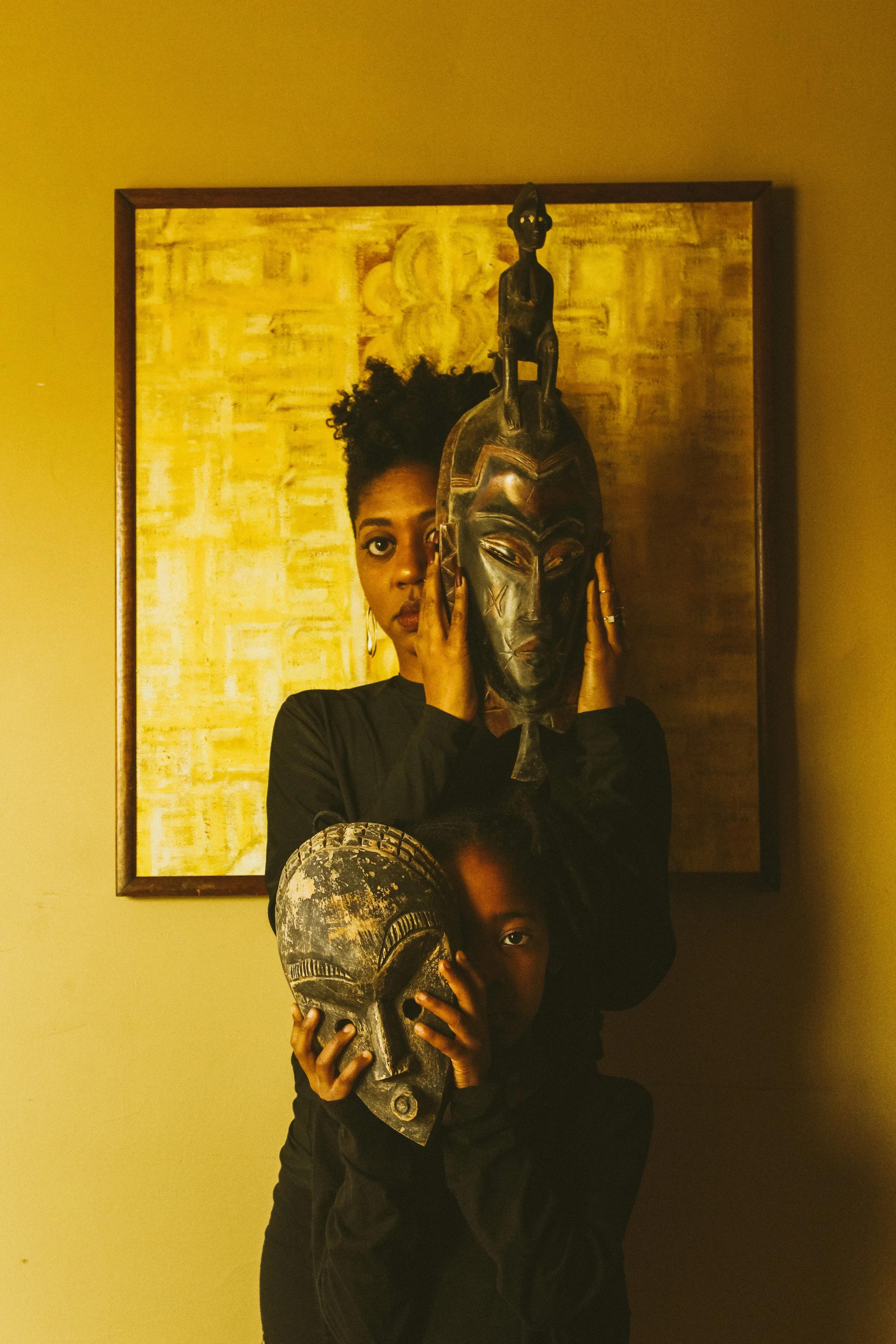 Two people, one woman and one child, holding African tribal masks in front of their faces, in a room with a framed yellow painting on the wall.