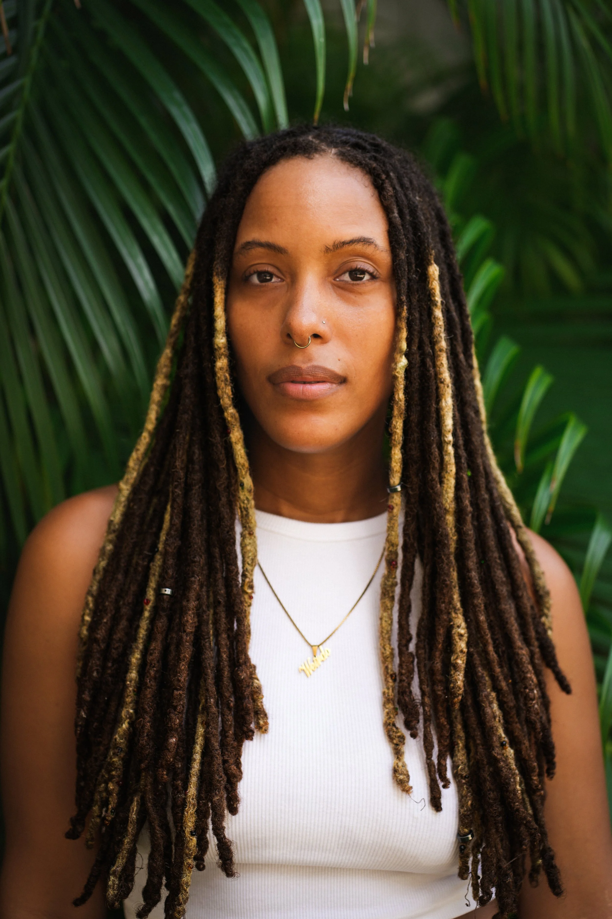 A woman with long dreadlocks and a nose piercing stands in front of green tropical plants, wearing a white sleeveless top and a necklace with a pendant that says 'Hello'.