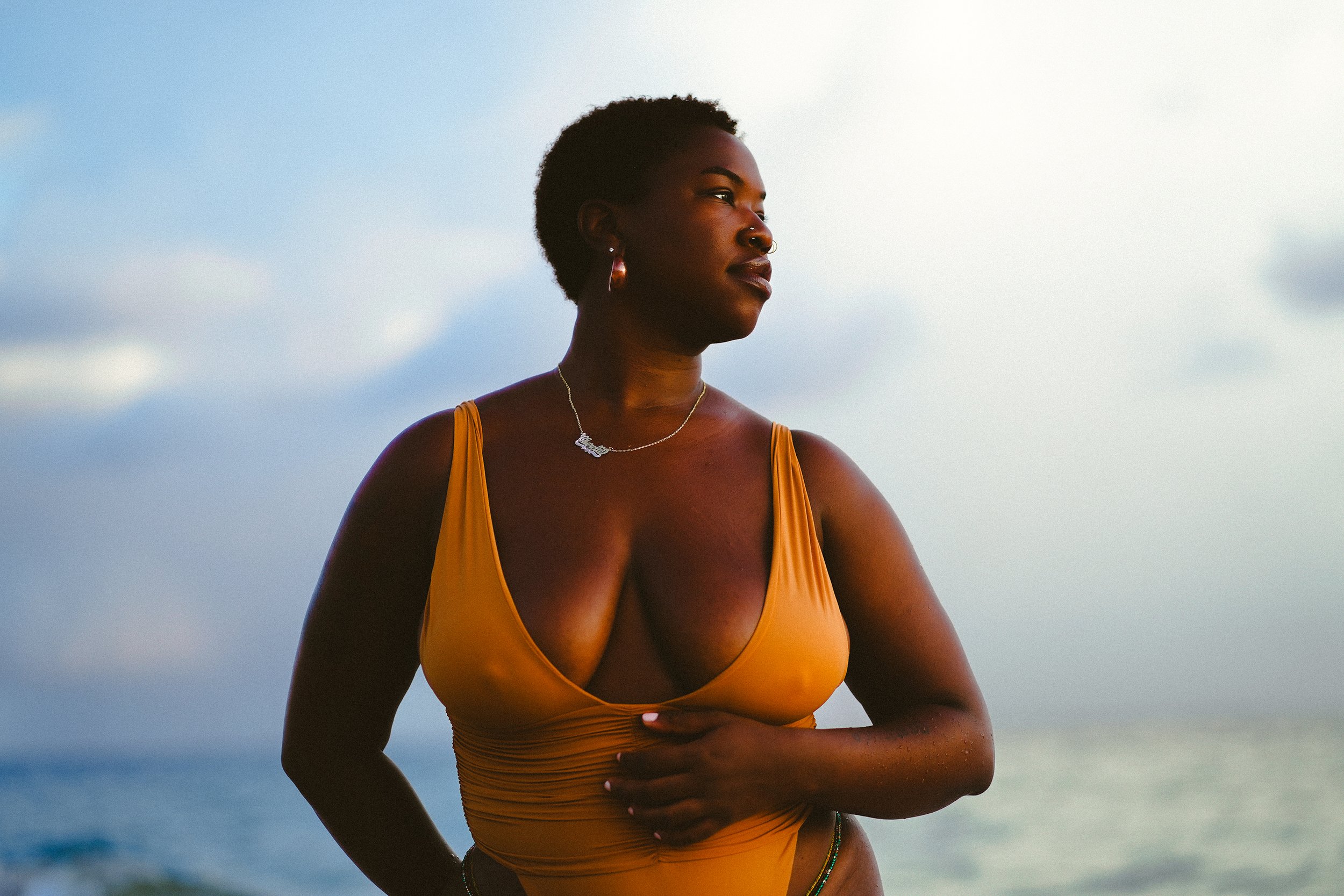 A woman with short curly hair and hoop earrings standing on a beach, wearing a yellow swimsuit, looking to the side with a calm expression, against a cloudy sky.