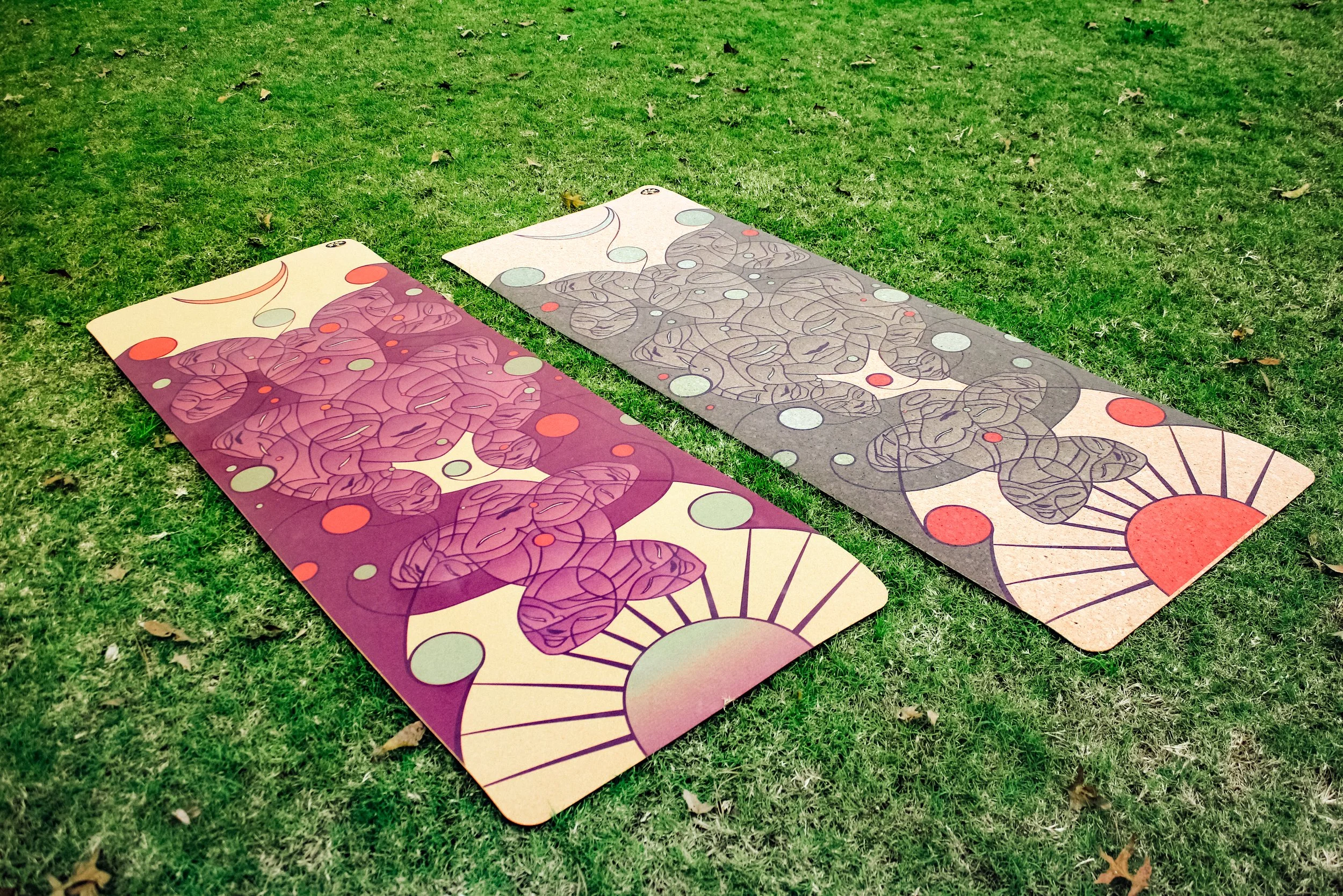 Two yoga mats laid out on green grass with scattered leaves, one purple with abstract designs and the other green with similar abstract patterns.