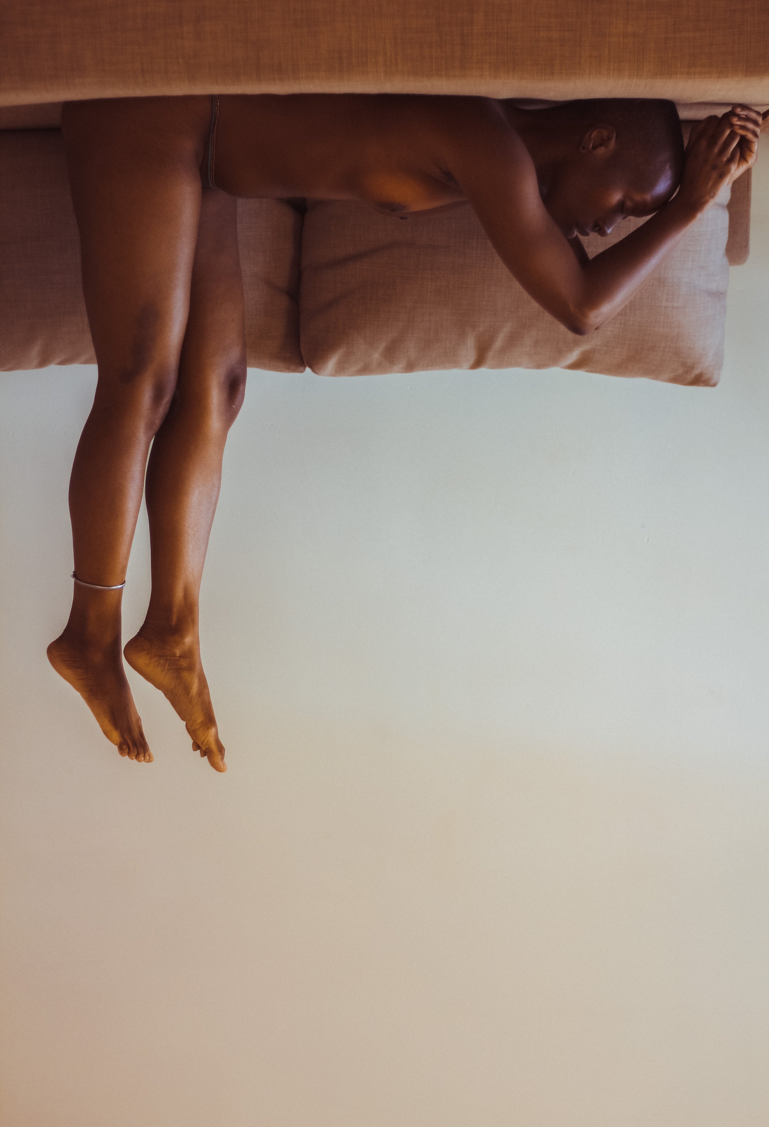 A man is lying on a couch with his eyes closed, holding his head with one hand. His body is nude except for a small tattoo on his thigh. The couch has a tan-colored cushion and the background is a plain wall.