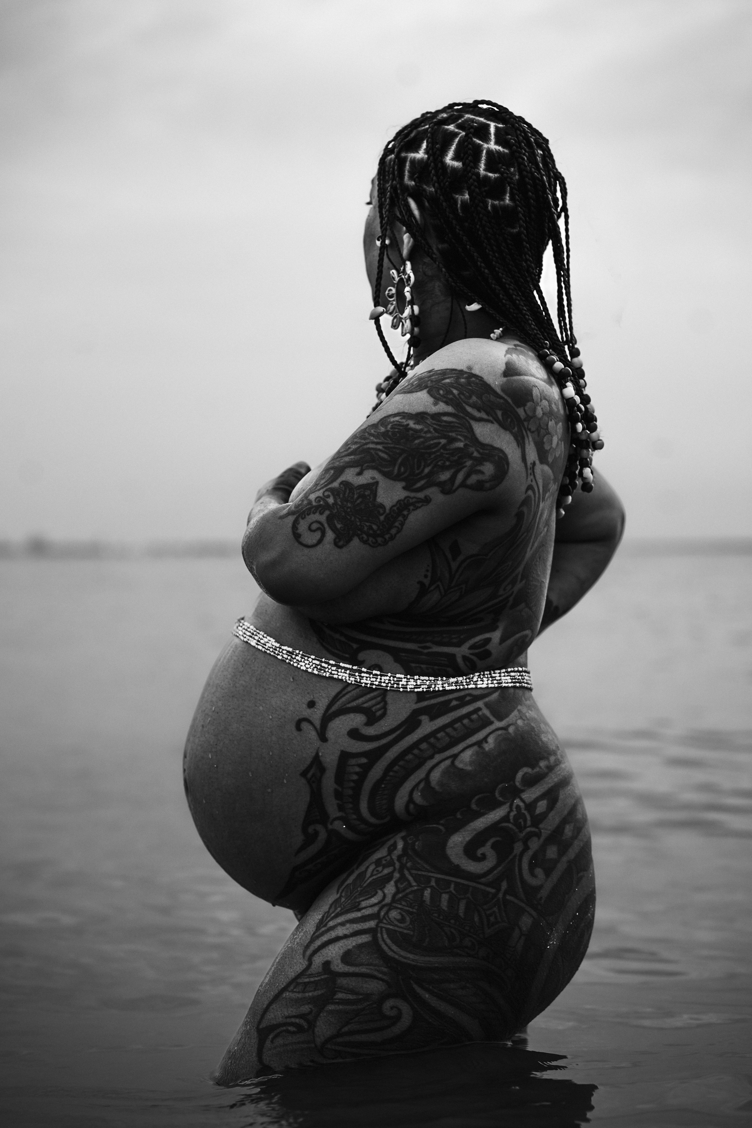 A woman with braided hair, tattoos on her arm and side, standing in the water at the beach, facing away from the camera.